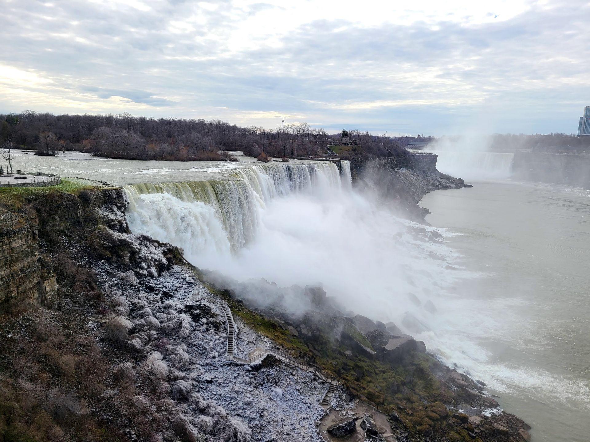 Looking at American Falls, a large waterfall. There is a light dusting of snow next to the water.