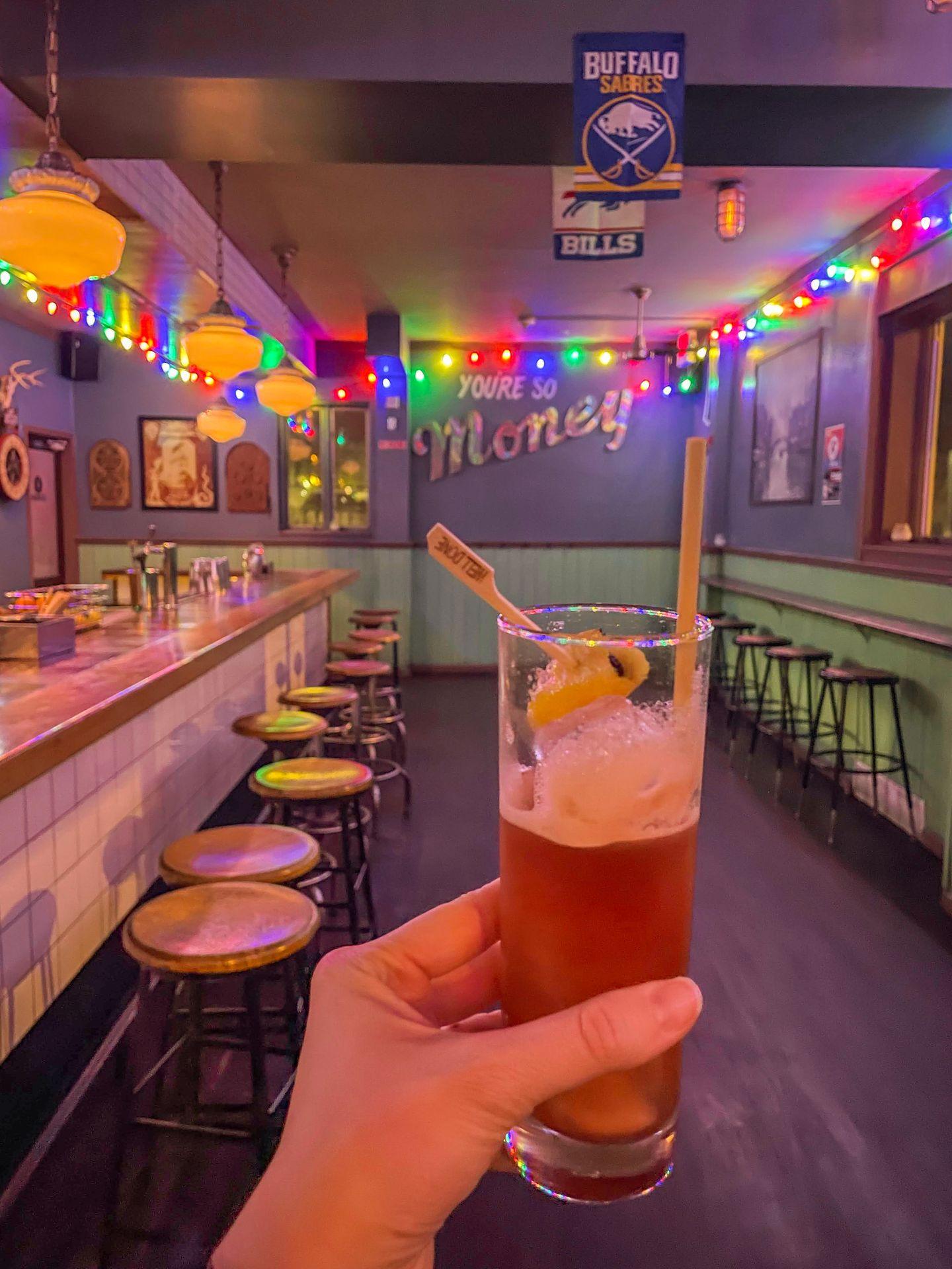 Holding up a cocktail inside of Ballyhoo. Colorful lights outline the corners of the walls and the wall reads 'You're So Money'