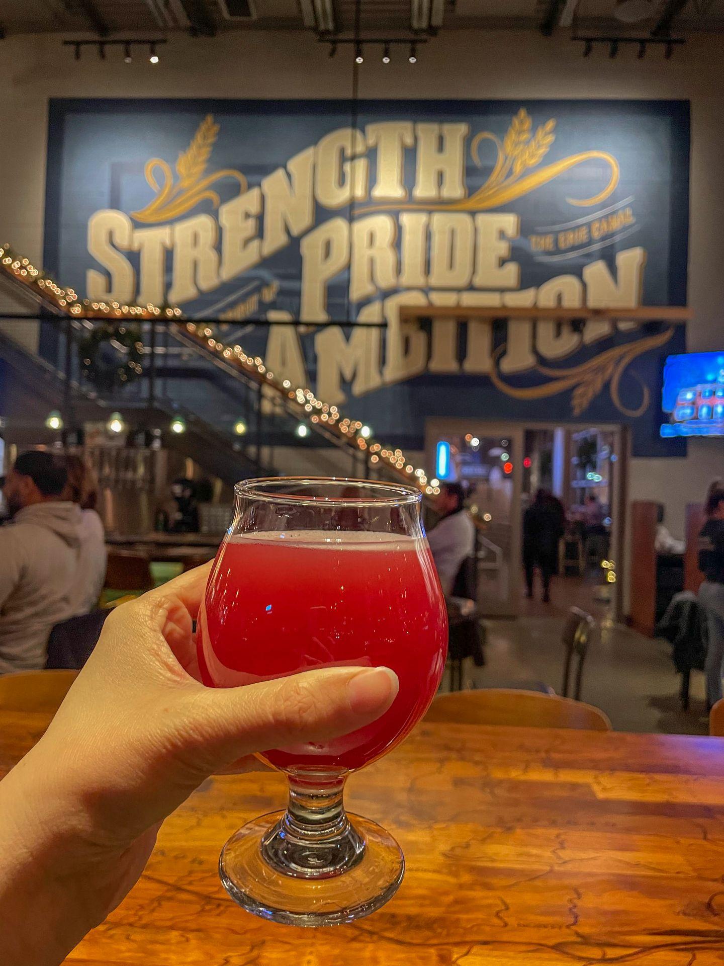 Holding up a glass with red beer. A mural in the background reads 'Strength, Pride, Ambition'