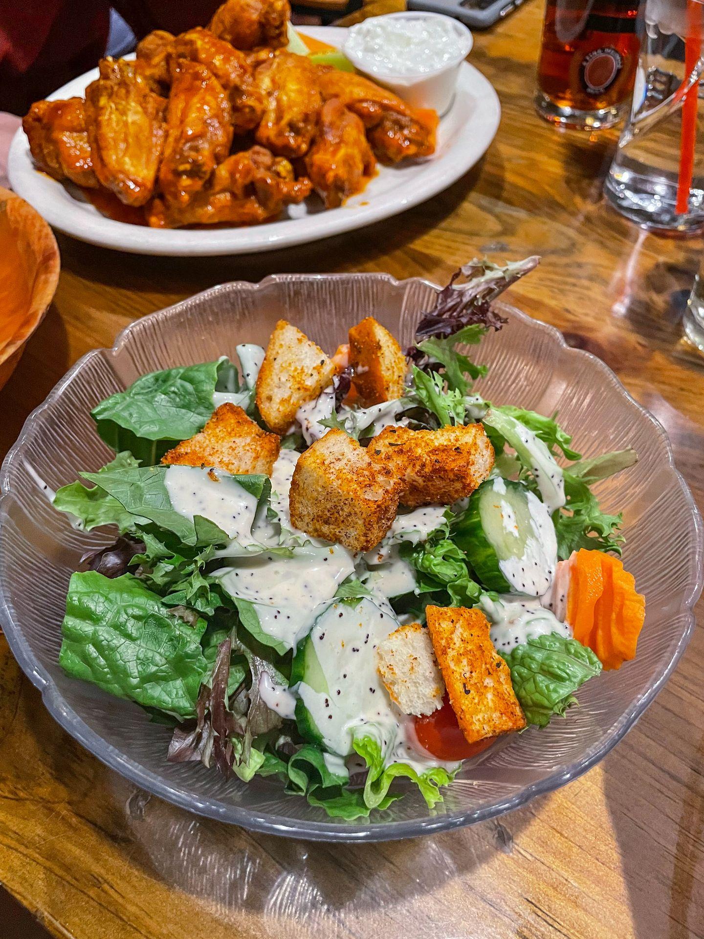 A salad with croutons and a plate of wings in the background.