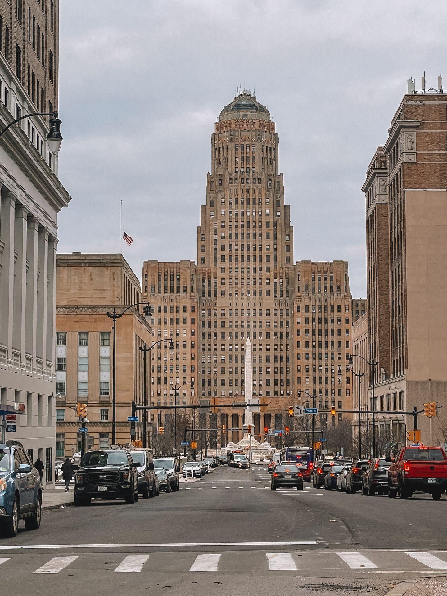Looking down the street at a view of Buffalo City Hall, a tall building that is light brown in color. A tall white pillar is outside of the building in the center.
