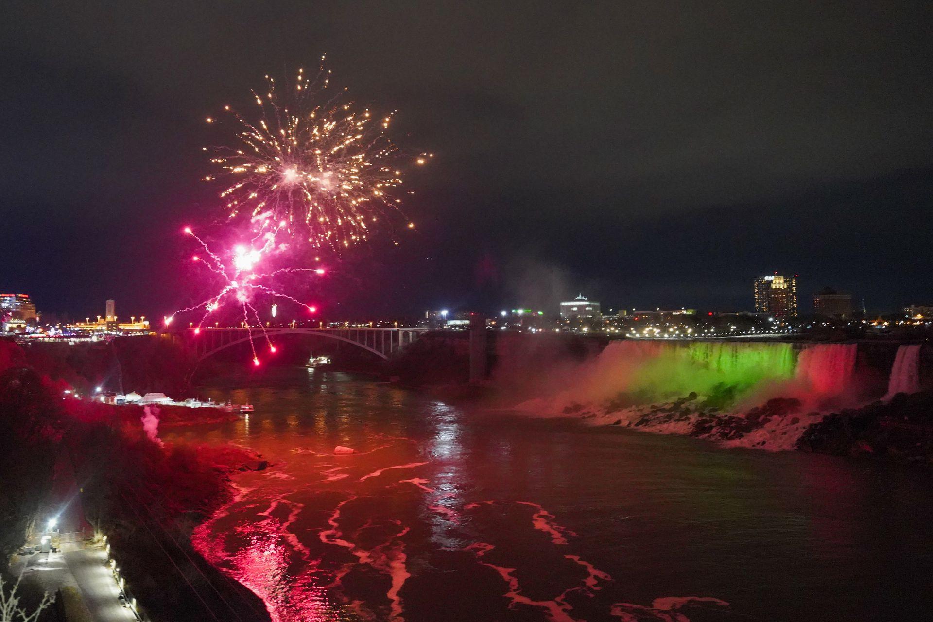 Fireworks over the Niagara River with American Falls lit up in green and red lights to the right.