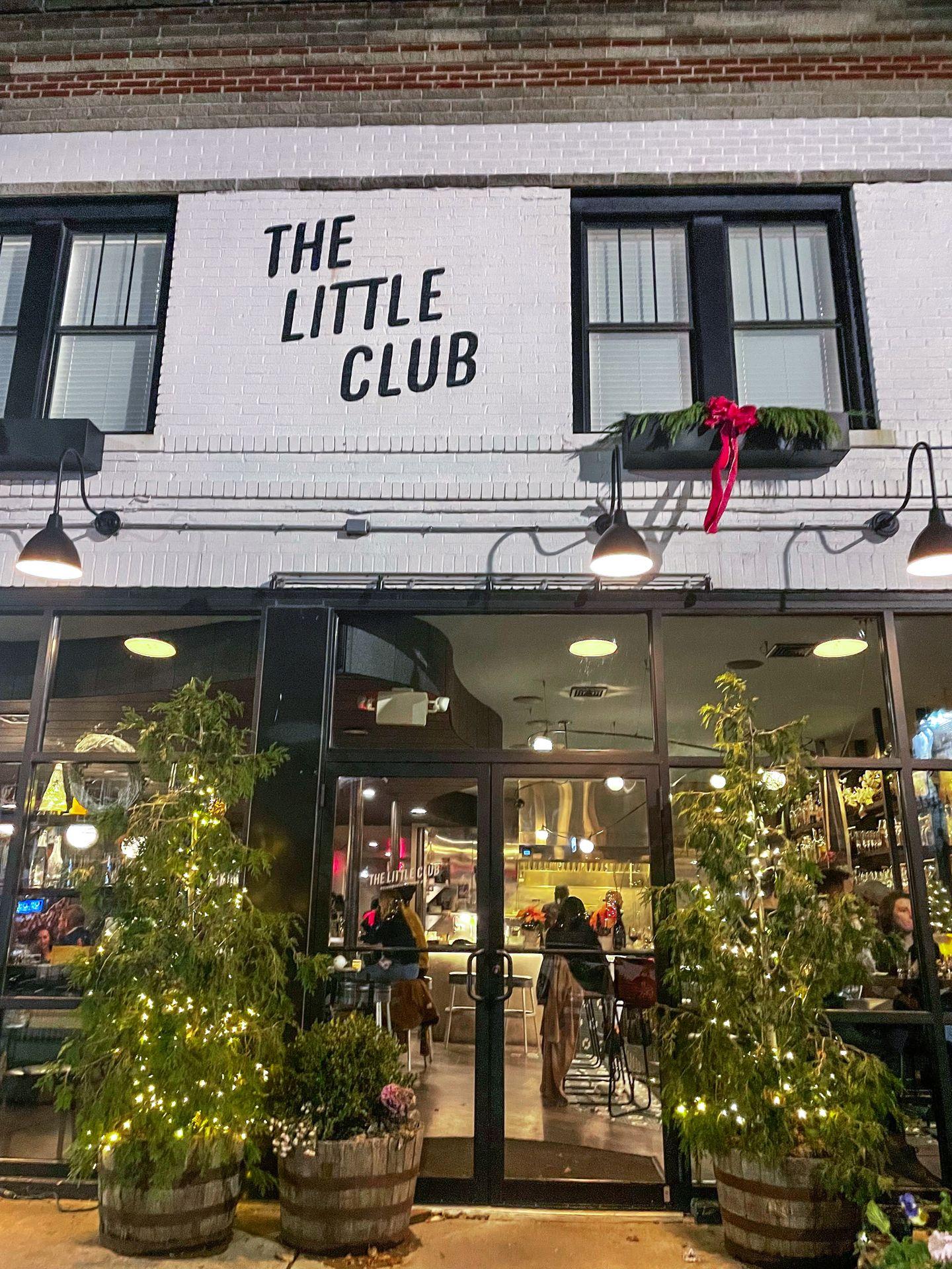 A white building that reads 'The Little Club.' There are Christmas trees outside of the doorway.