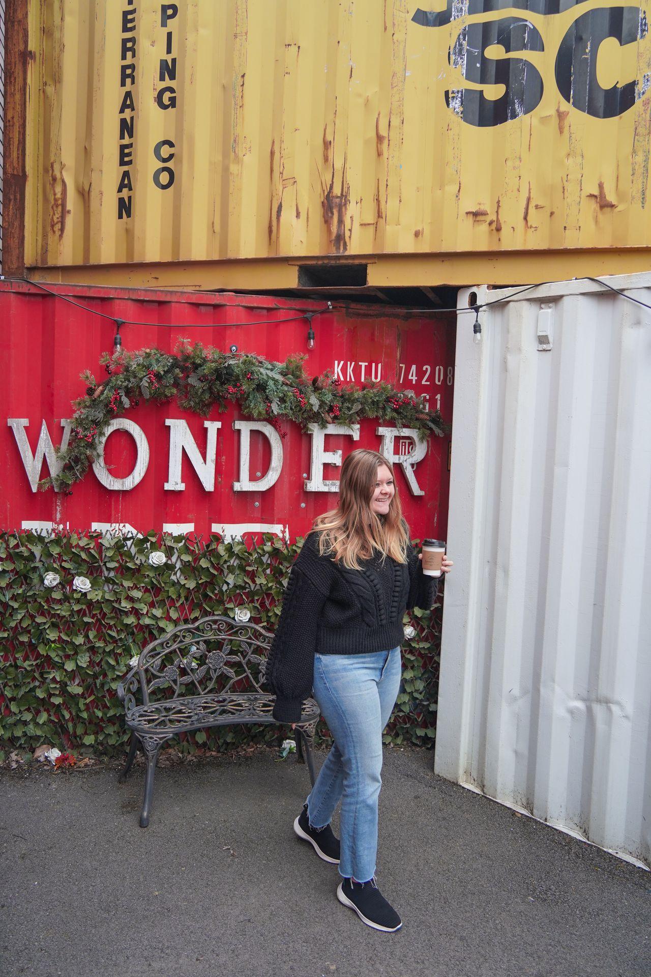 Lydia holding a coffee and standing in front of a display with 3 shipping containers, which are yellow, red and white. The red one reads 'Wonder' and is partially covered with greenery.