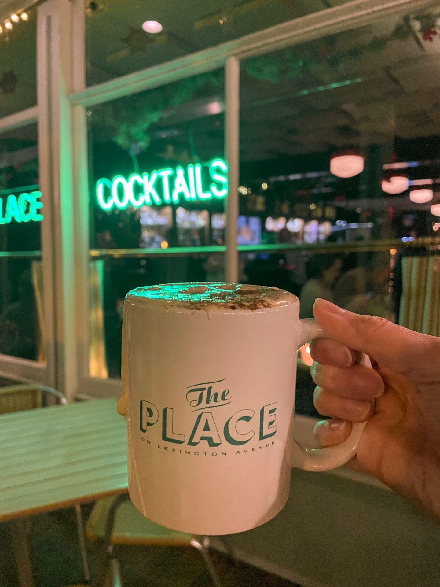 Holding up a mug full of an eggnog cocktail. The white mug reads 'The Place' in green letters.