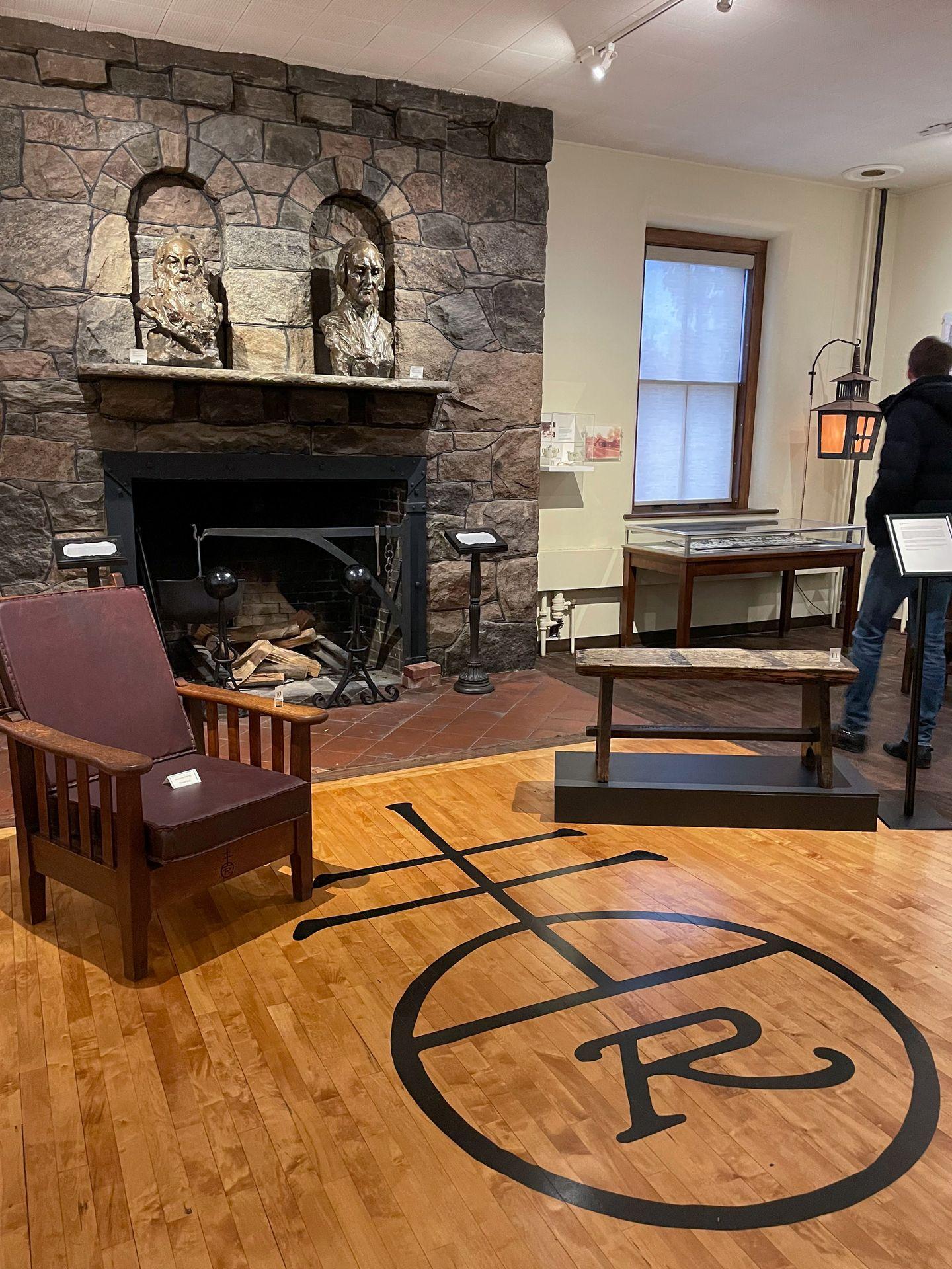 A room with a stone fireplace, a chair and a symbol on the floor for Roycroft furniture.