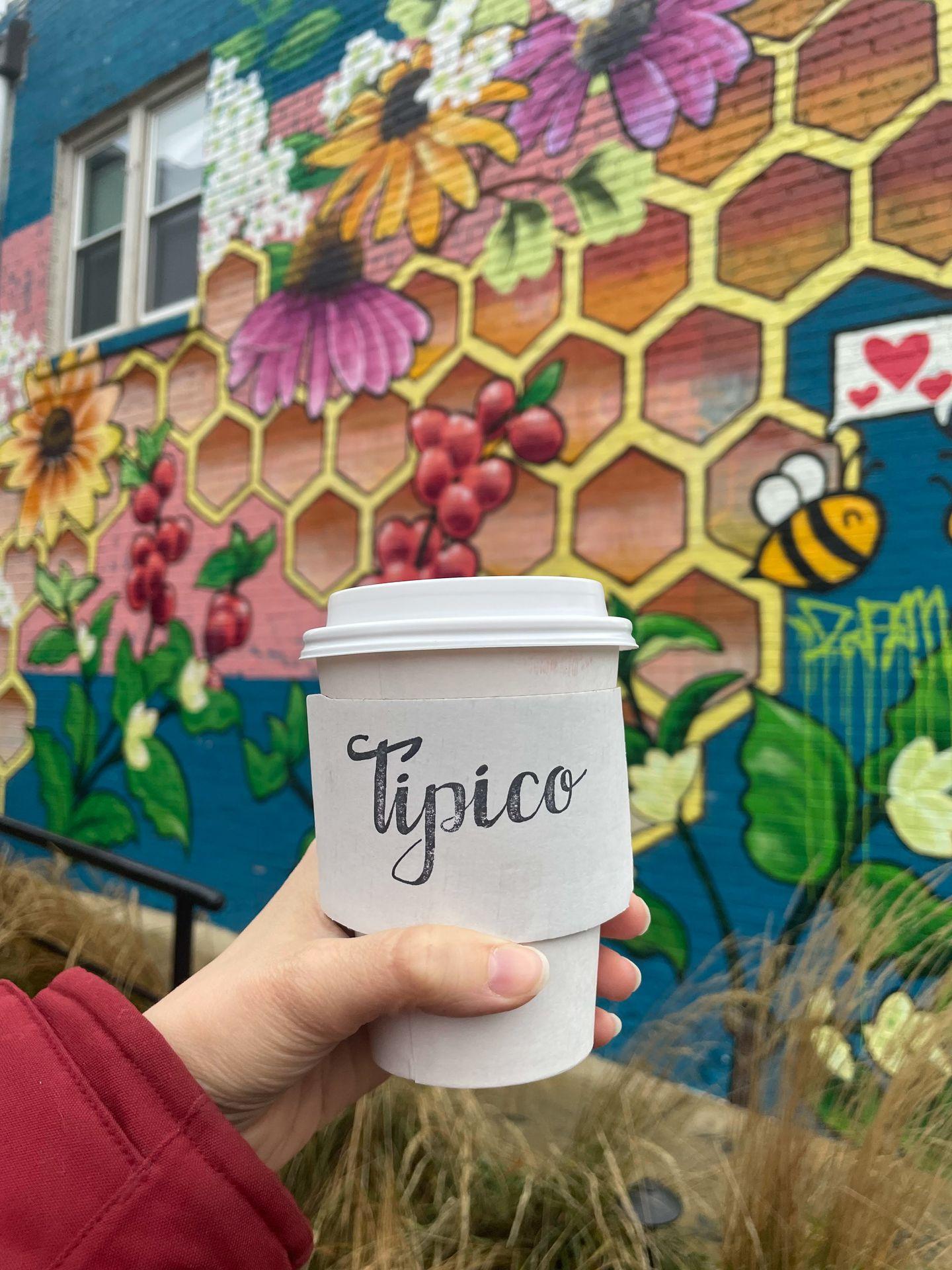 Holding up a white coffee to go mug that reads 'Tipico.' There is a colorful mural in the background that depicts a honeycomb, a bee and flowers.