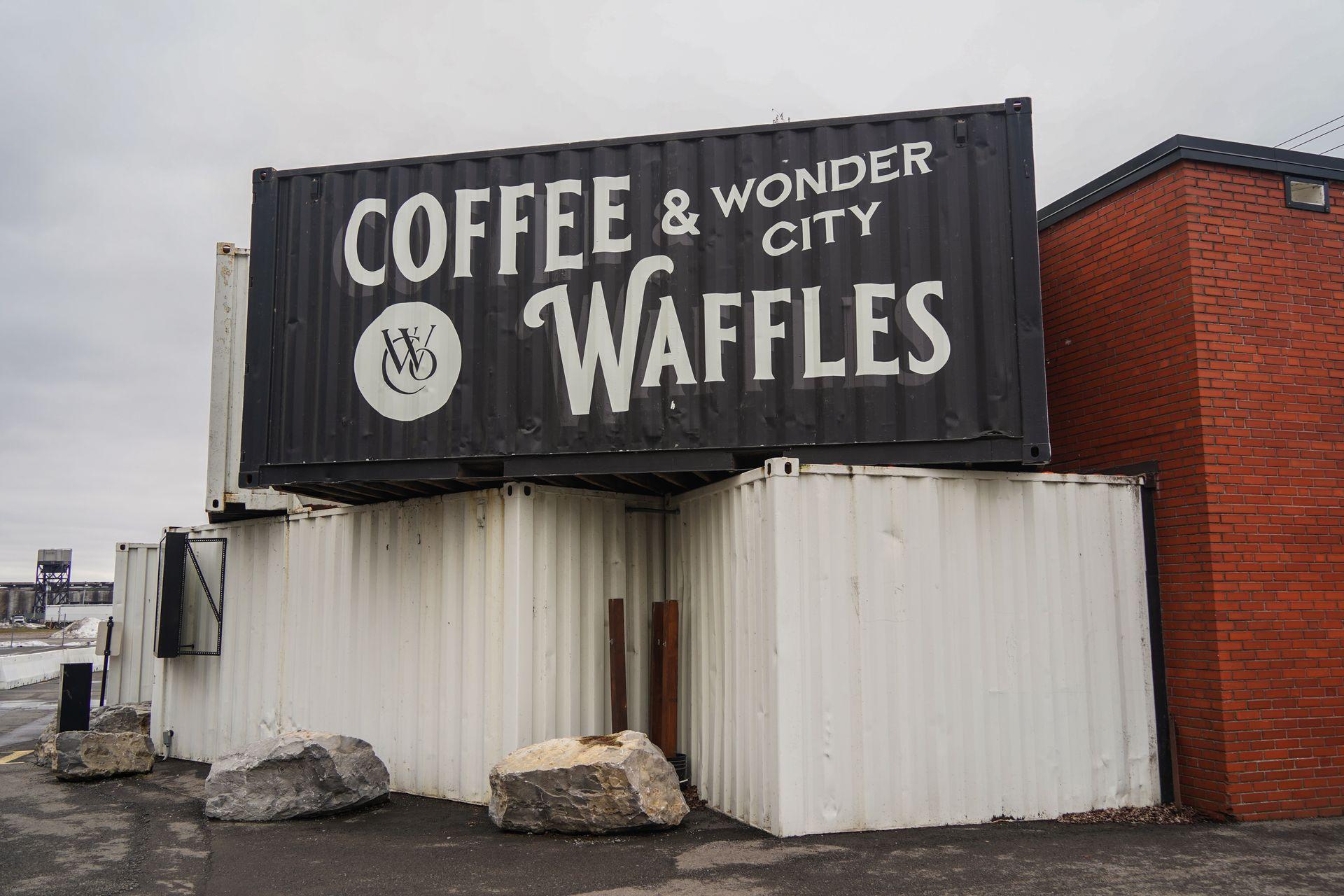 A black shipping container stacked on top of white shipping containers. The black one reads 'Coffee & Waffles Wonder City'