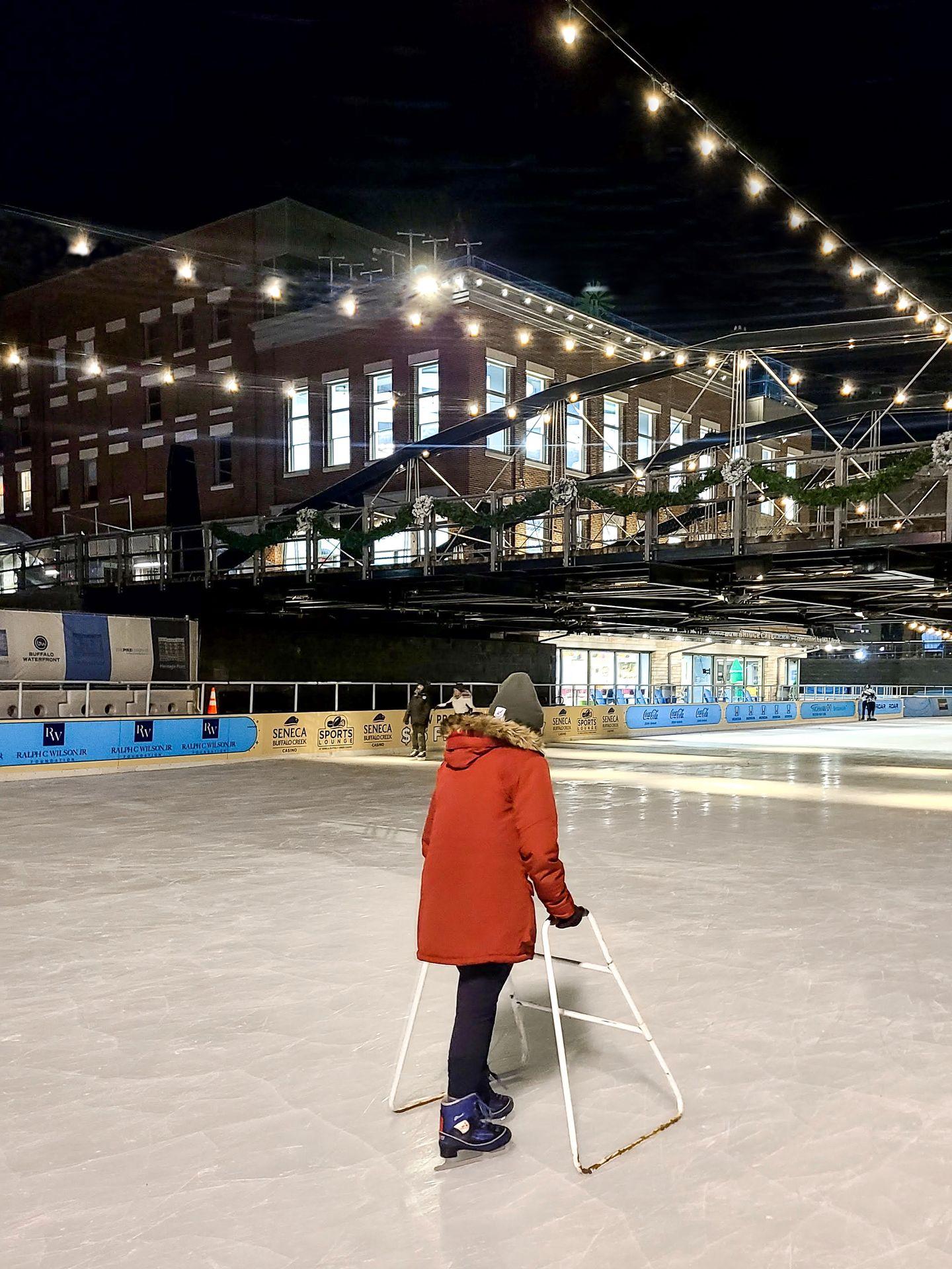 Lydia ice skating using a walker. There are hanging lights and a bridge above the rink. Lydia wears a red coat and green hat.