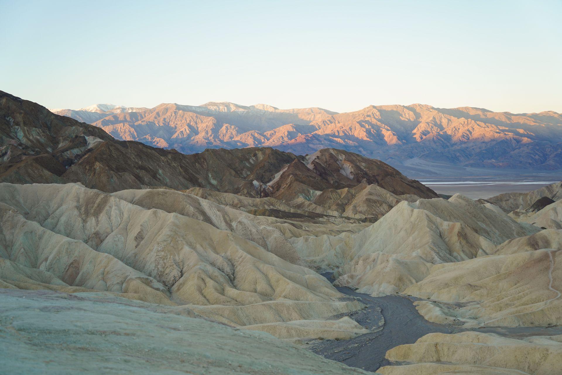 A landscape with badlands in various shades of brown. In the distance, mountains glow pink from the rising sun.