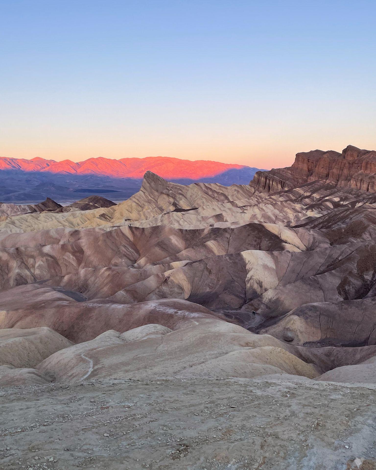 A scene with badlands that look to be striped in various shades of brown. The mountains in the distance glow pink and blue from the rising sun.