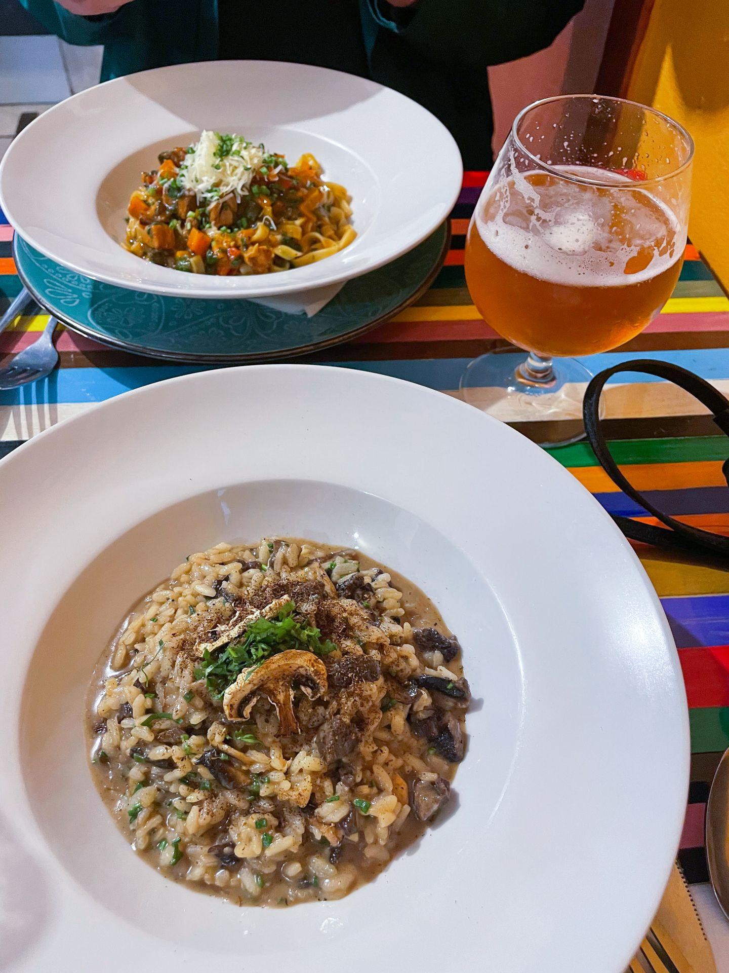 A large white bowl of mushroom risotto and another bowl of pasta across the table. There is also a large glass of beer.
