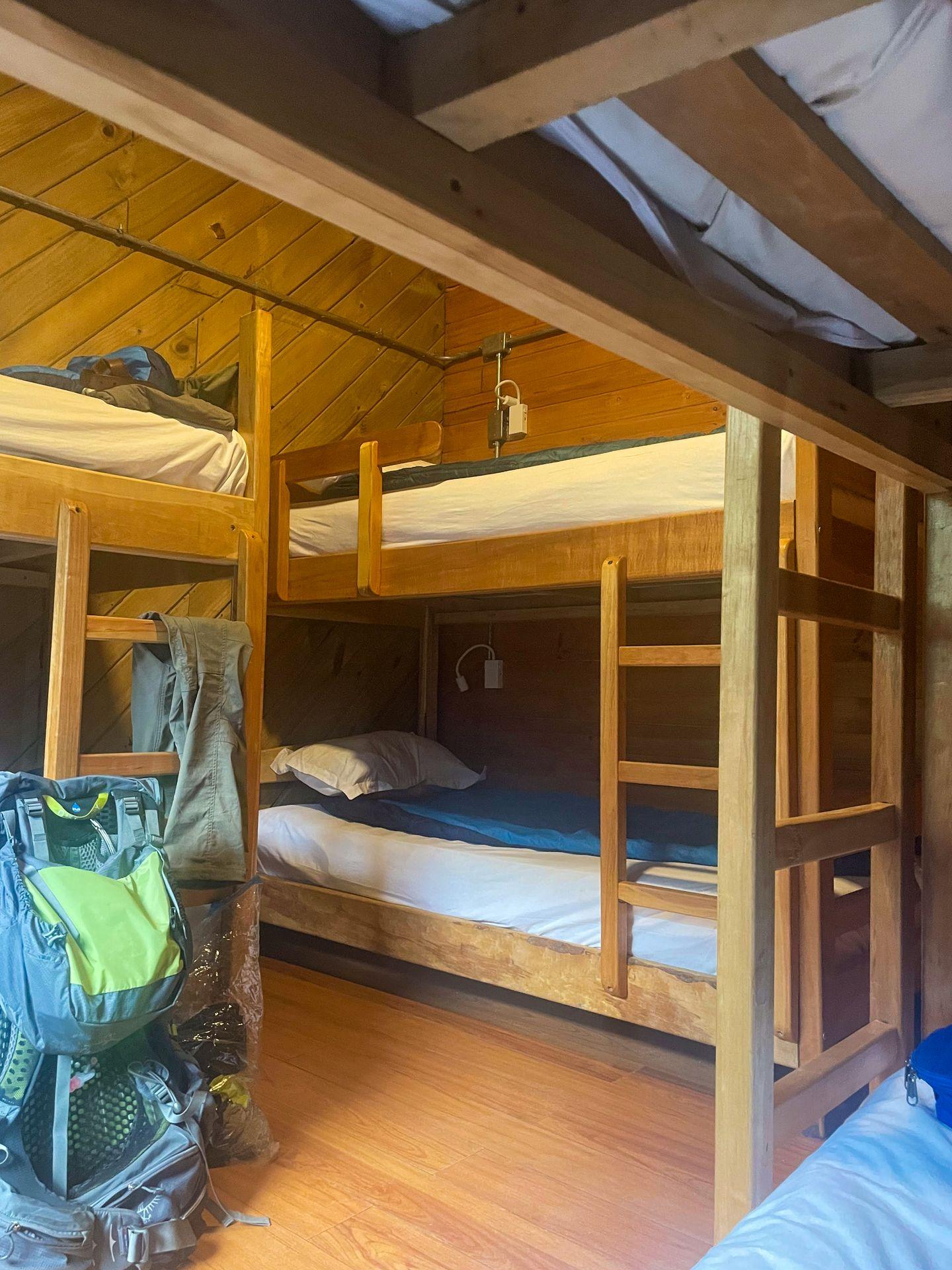 A room with 3 bunkbeds and a large backpack on the floor. Sleeping bags lay across the mattresses.