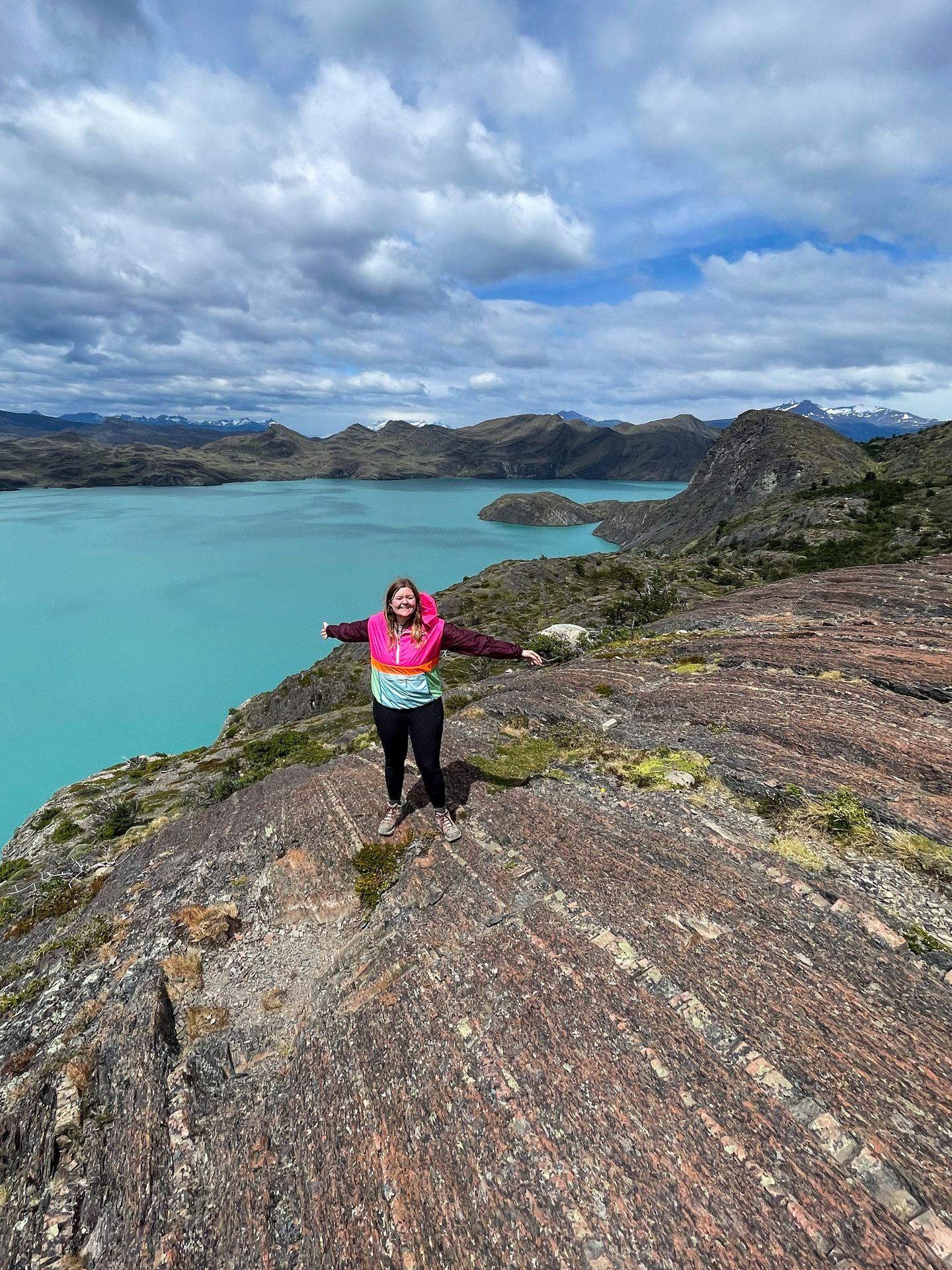 Lydia standing on a curved rockface with her arms in the air. In the background, there is a lake with a brilliant blue color.