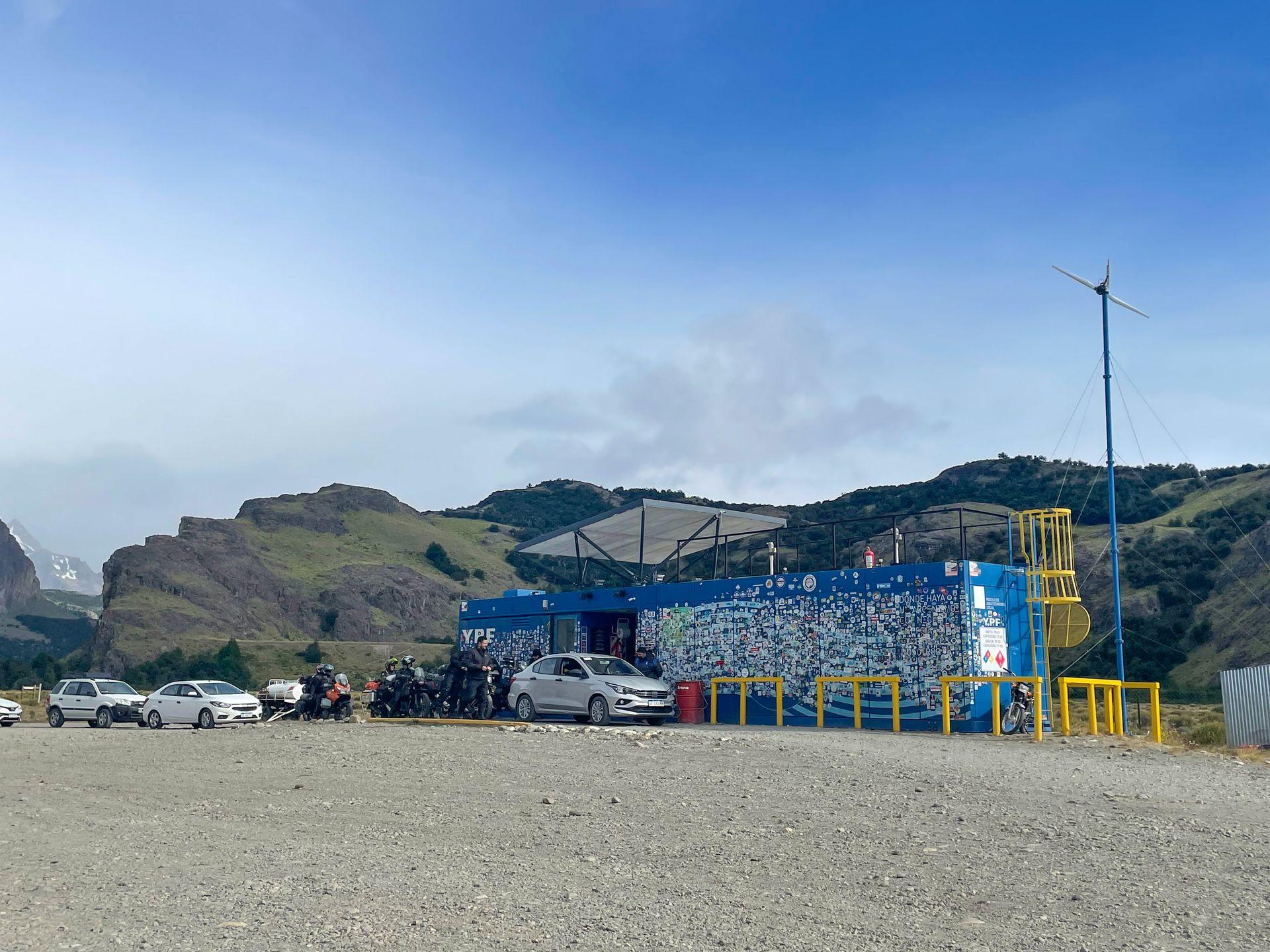 A line of cars and motorcycles at the one gas station in El Chalten. The gas station is a small blue building covered in stickers.