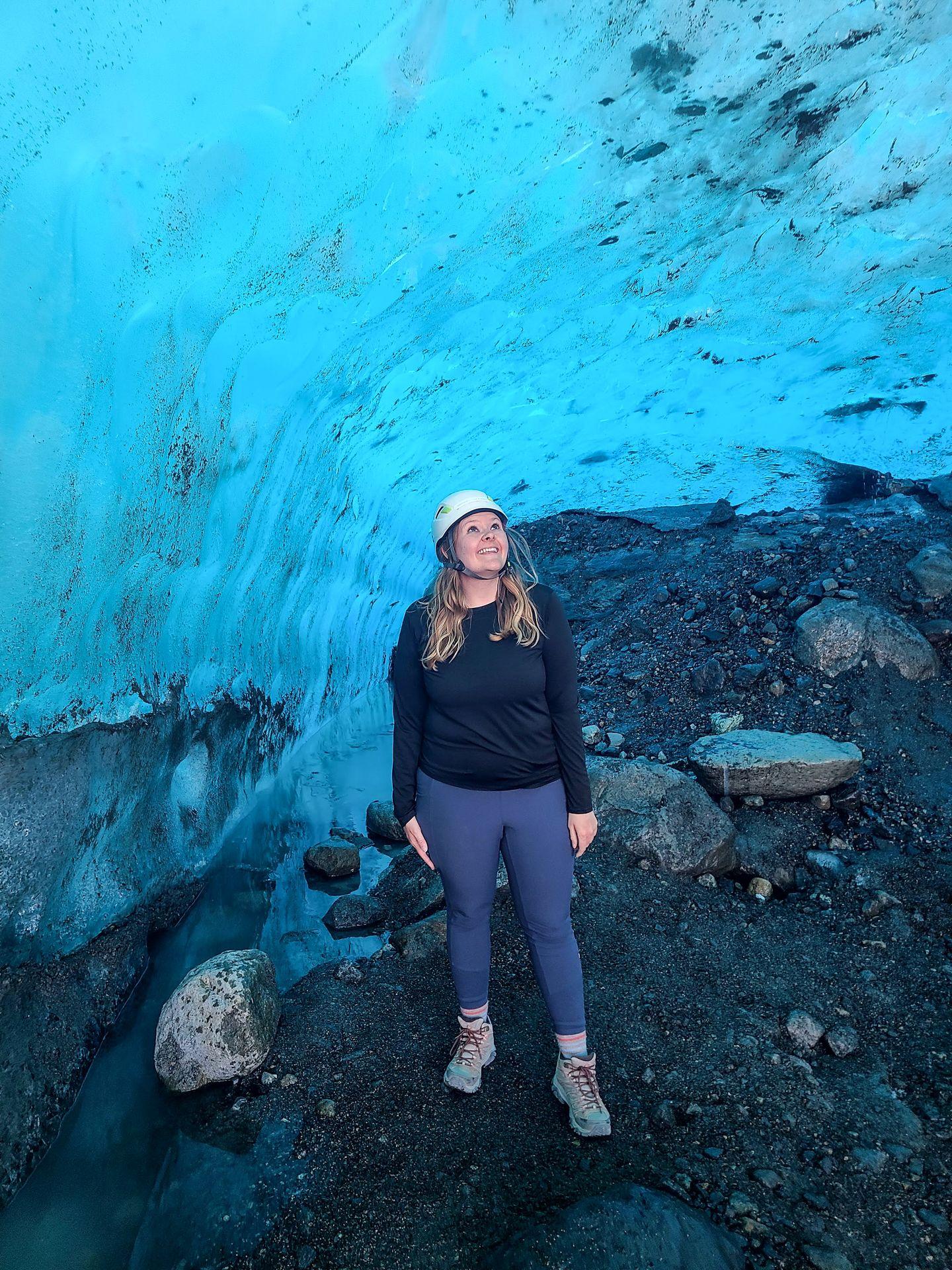 Lydia standing inside of an ice cave made of bright blue ice. She wears a helmet, blue leggings and a black shirt.