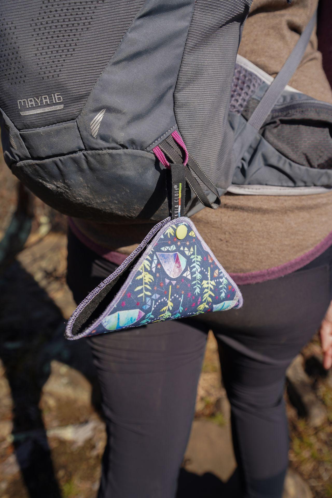 A kula cloth hanging from a hiking backpack. It is clipped onto a loop and shaped like a taco.