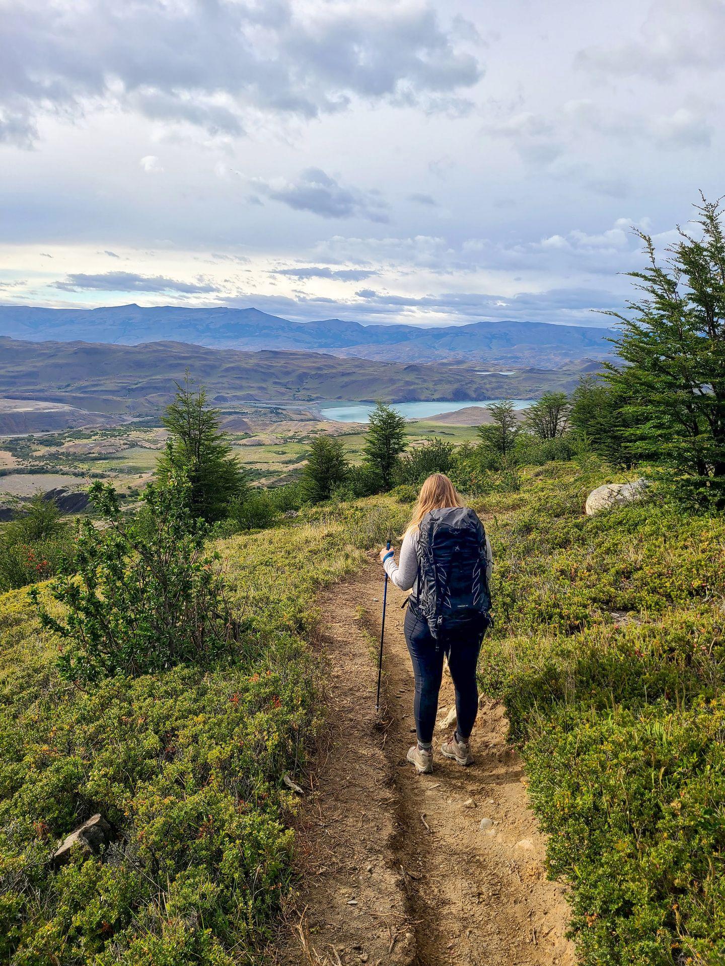 Lydia standing on a trail in the W Trek wearing a black backpack. The trail is surrounded by greenery and there is a lake in the distance.