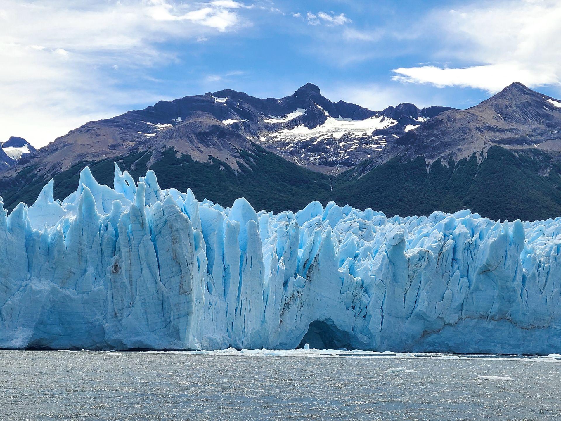 A giant blue glacier on the edge of the water with mountains in the background. The glacier is a brilliant blue and full of jagged ice formations.