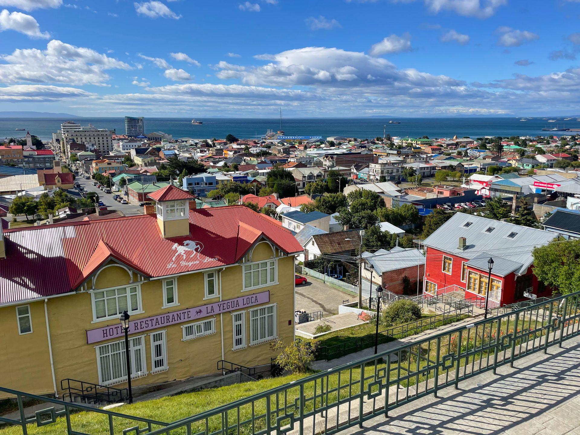 Looking down at a colorful city with the ocean in the distance. In the foreground is a yellow hotel with a red roof.