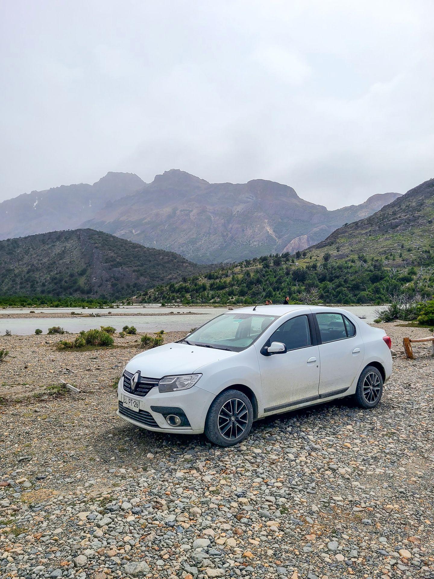 A small white car next to a lake with mountains in the background.