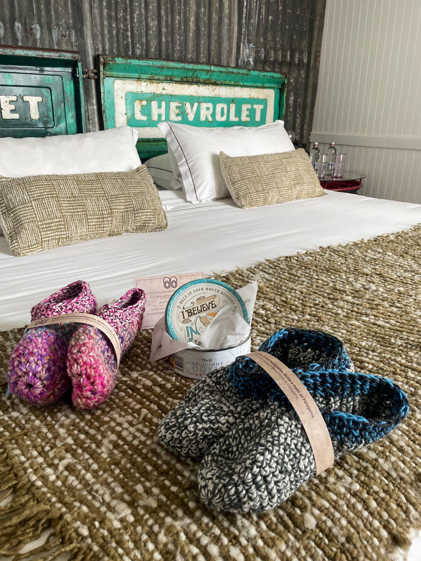 Two pairs of slipper and a tin of chocolate sitting on a bed. Behind the pillows is a piece of vintage sign that reads 'Chevrolet'