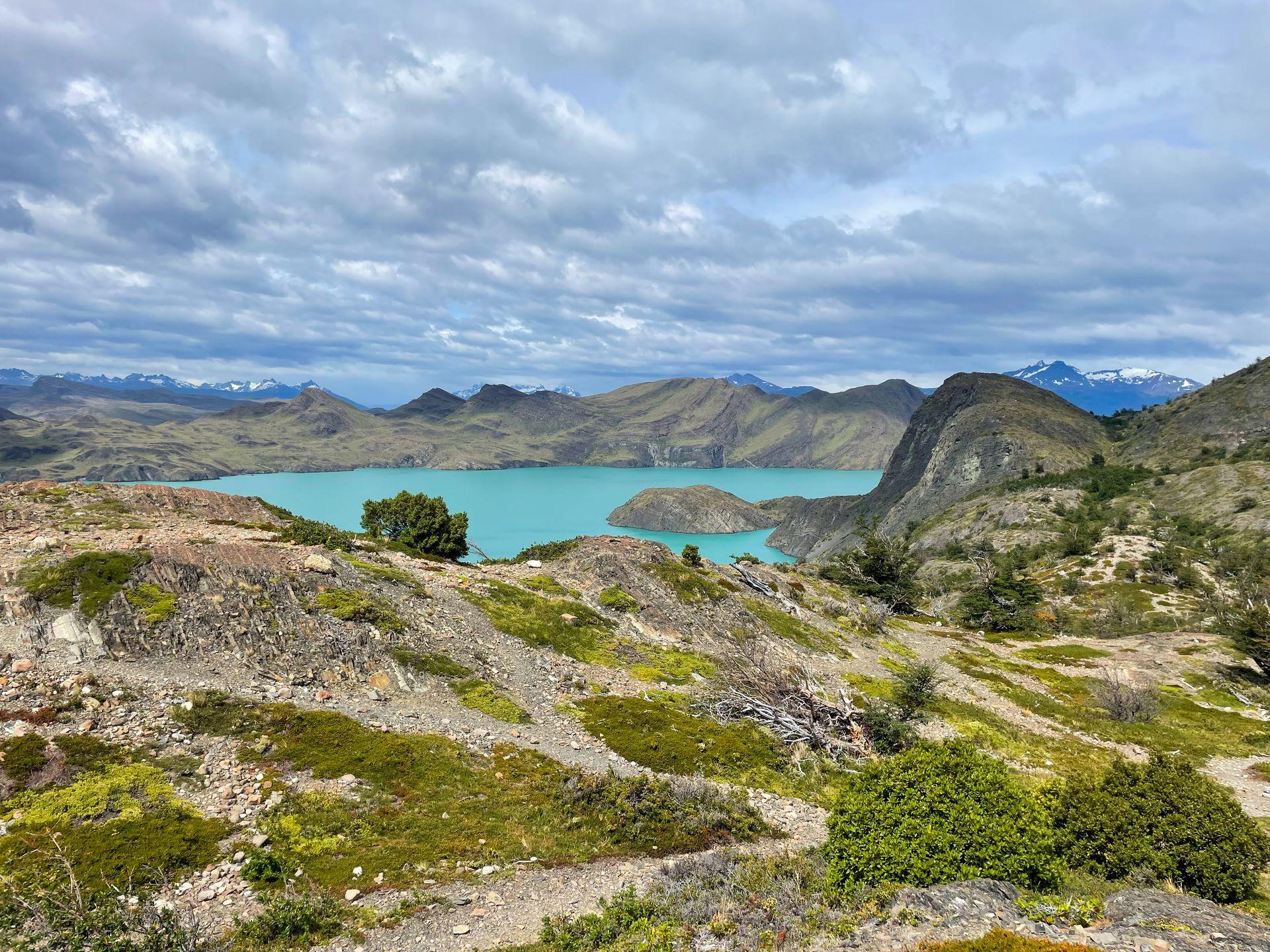 A bright blue lake surrounded by mountains. There is a mix of greenery and rocky areas across the mountainous landscape.