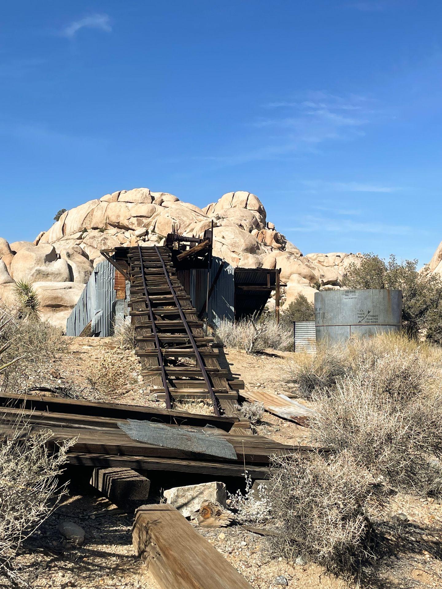 Former mining ruins that include a tilted wooden chute and silver structures. There is a pile of tan boulders in the background.