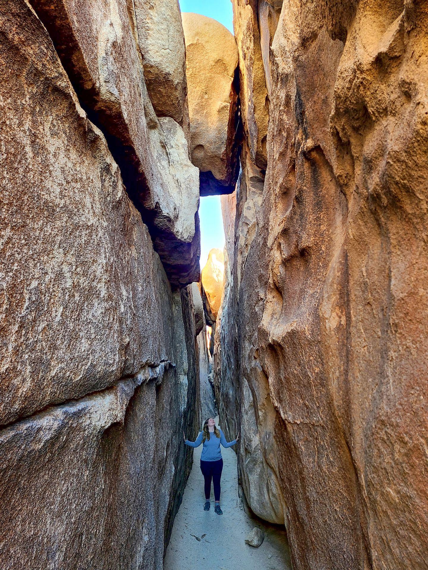 Lydia standing in a towering slot canyon and looking up towards the opening above. A giant boulder sits on top of the canyon.