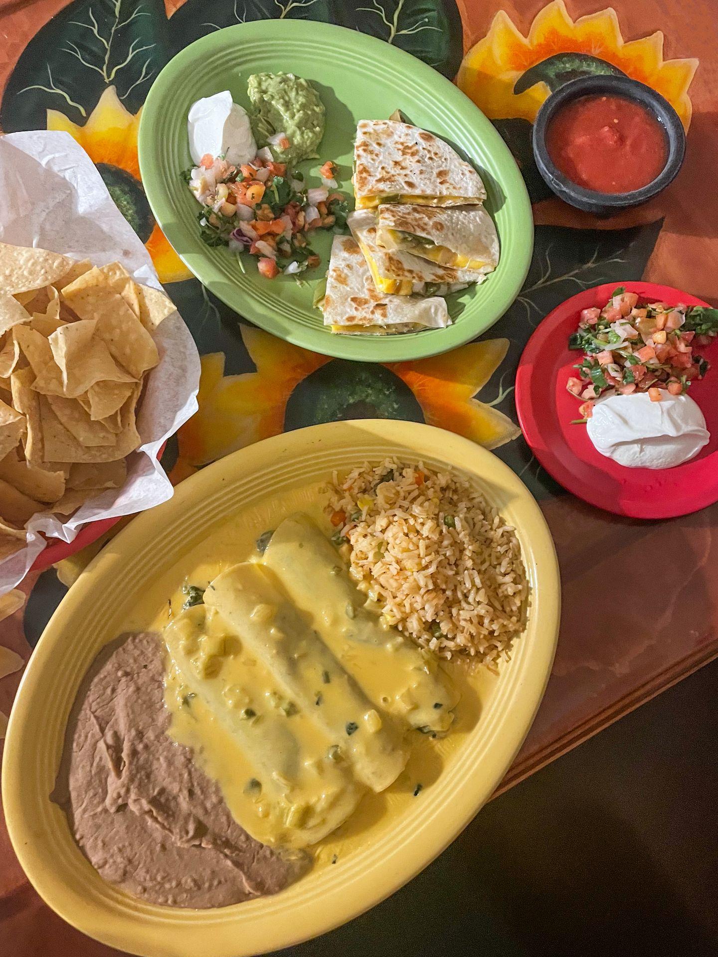 A couple colorful plates of Mexican food, next to a bowl of chips,  at Carmela's. There are enchilada's served with rice and beans and quesadillas served with guacamole, sour cream and pico de galo.