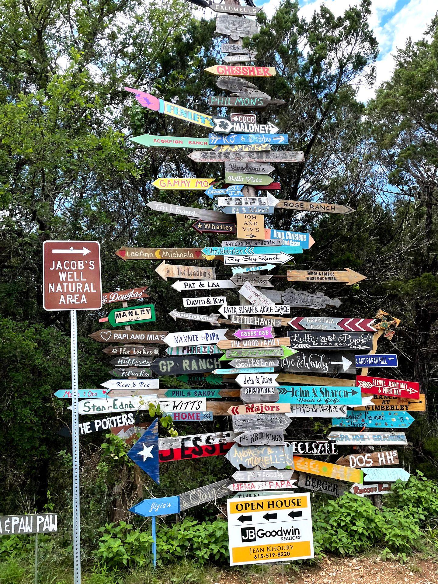 A tall sign post 100's of arrows with various family names around the area. There is also a brown sign that reads 'Jacob's Well Natural Area'