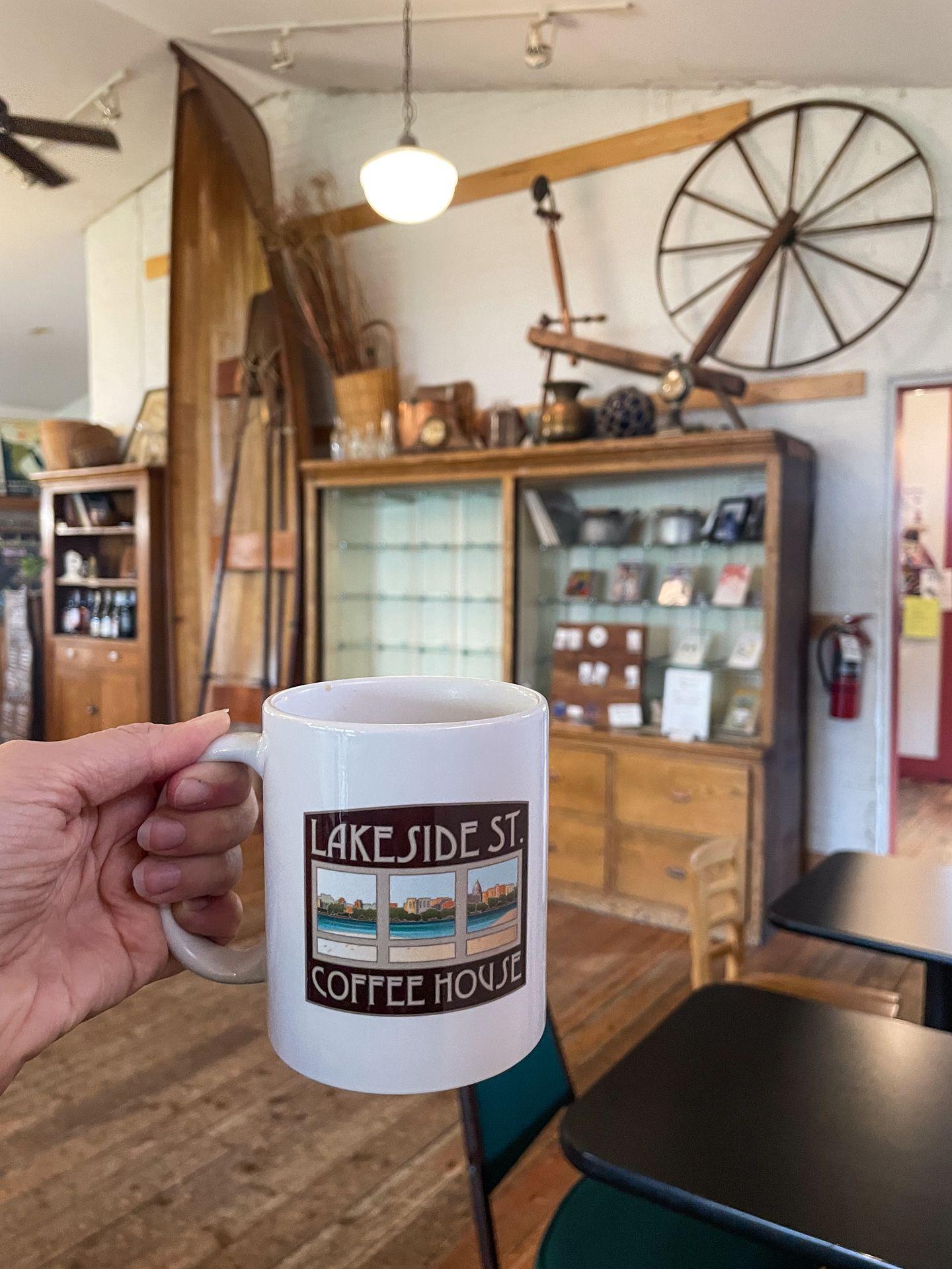 Holding up a coffee mug at Lakeside Coffee. The mug reads 'Lakeside St Coffee House' and has an artistic rendering of the windows in the coffee shop.