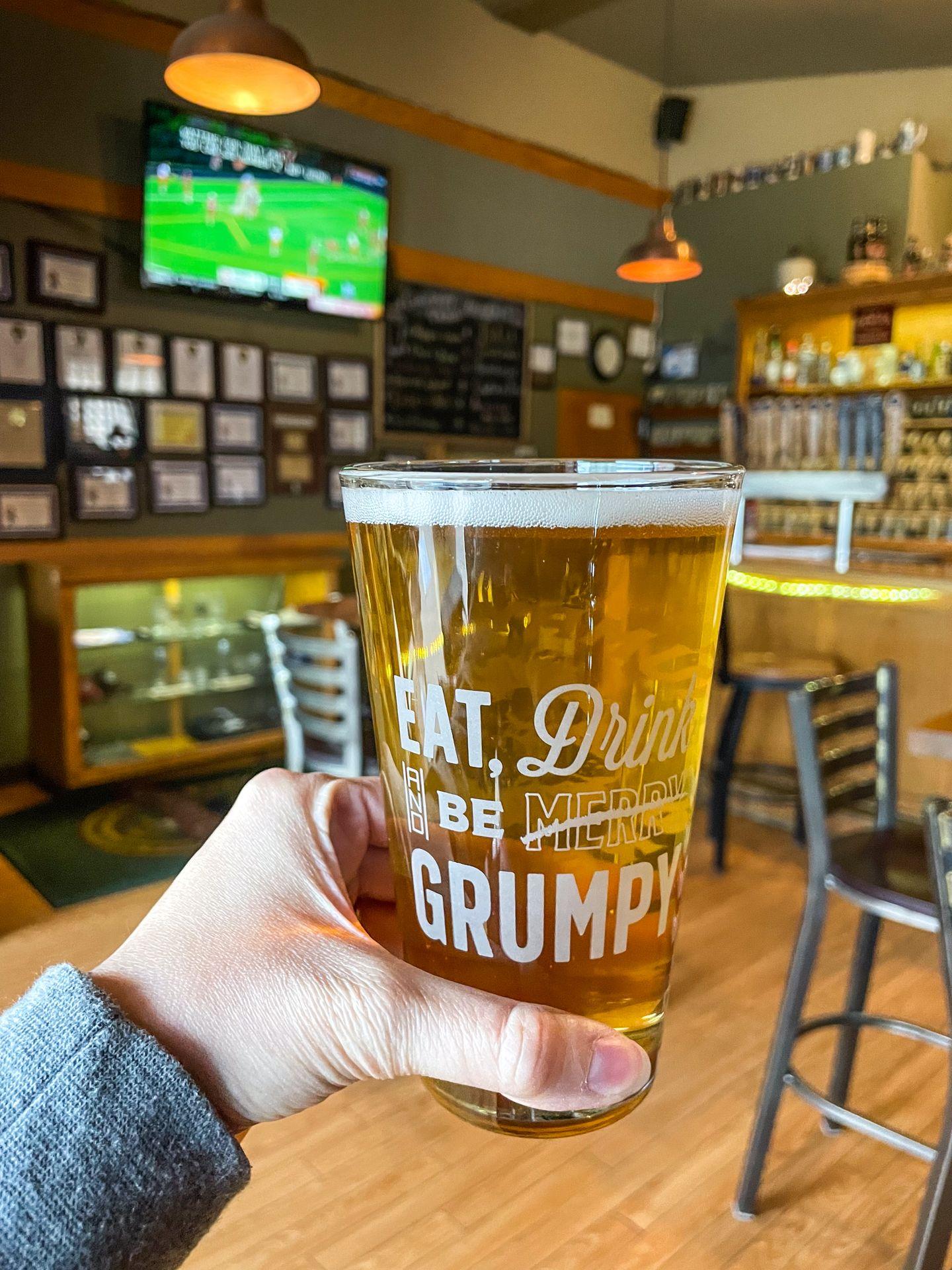 A glass of beer from Grumpy Troll Brew Pub. The glass reads "Eat, Drink, and Be Merry" but "Merry" is crossed out and replaced with Grumpy