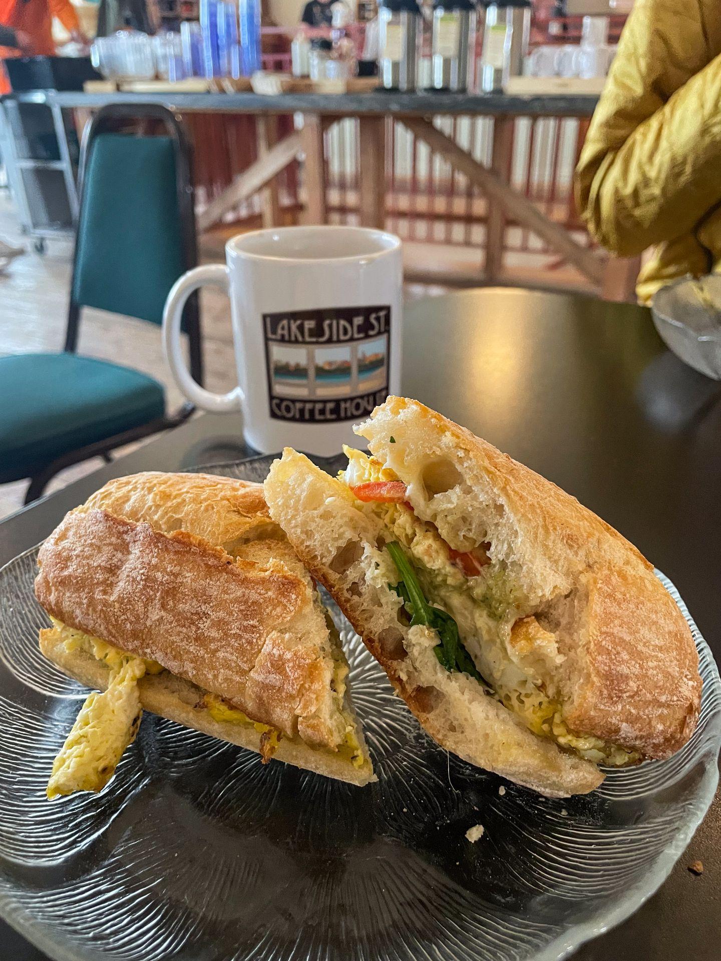 A sandwich on ciabatta bread with a mug behind it sitting on the table at Lakeside Coffee.