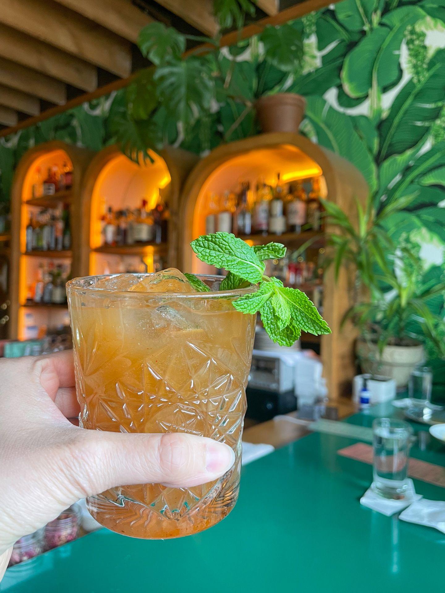 Holding up a cocktail at the bar at Mint Mark. The walls behind the bar are decorated to look like greenery.
