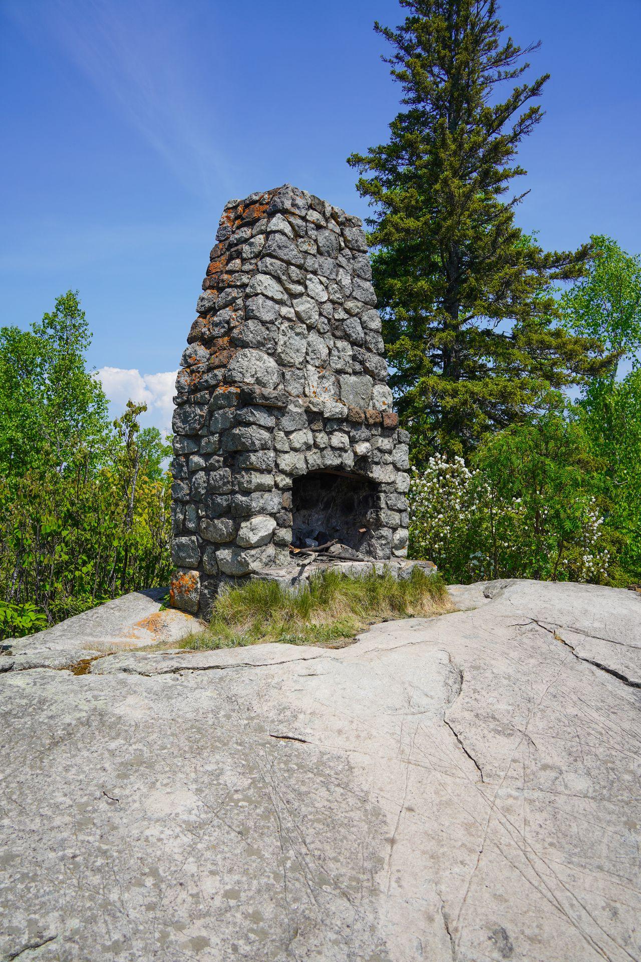 A stone fireplace stands on it's own, surrounded by rock and some trees.