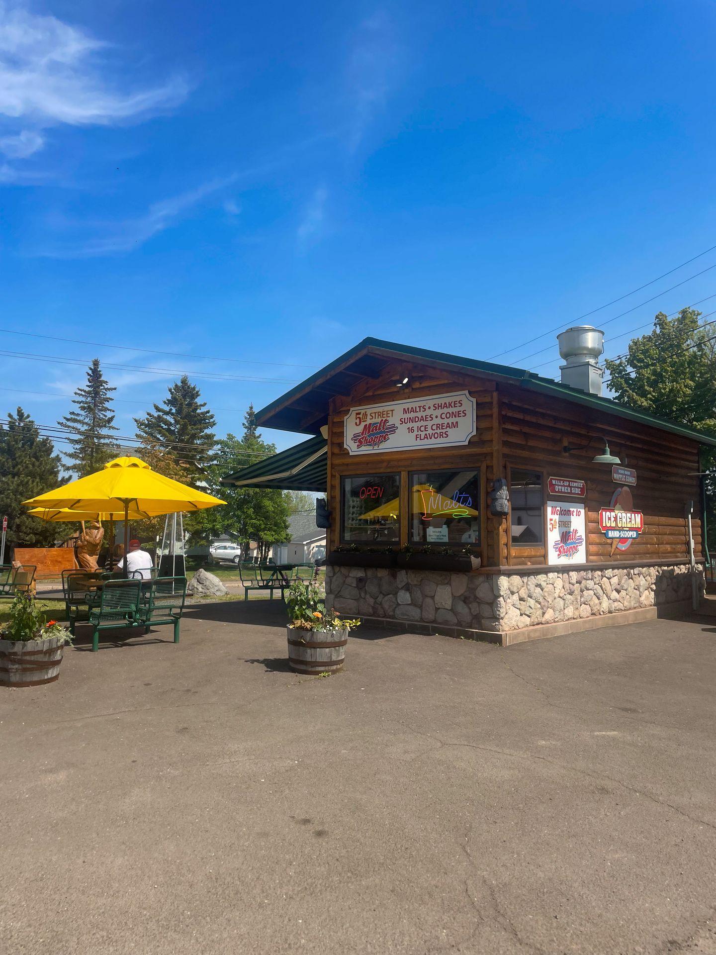 The exterior of 5th Street Malt Shoppe, a small roadside building that looks like it's made of logs. There is a table with a yellow umbrella in front of the building.