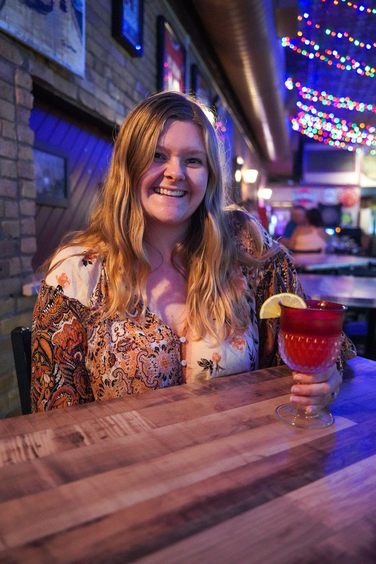 Lydia holding a strawberry margarita cocktail as she sits at a table inside the Pickled Loon