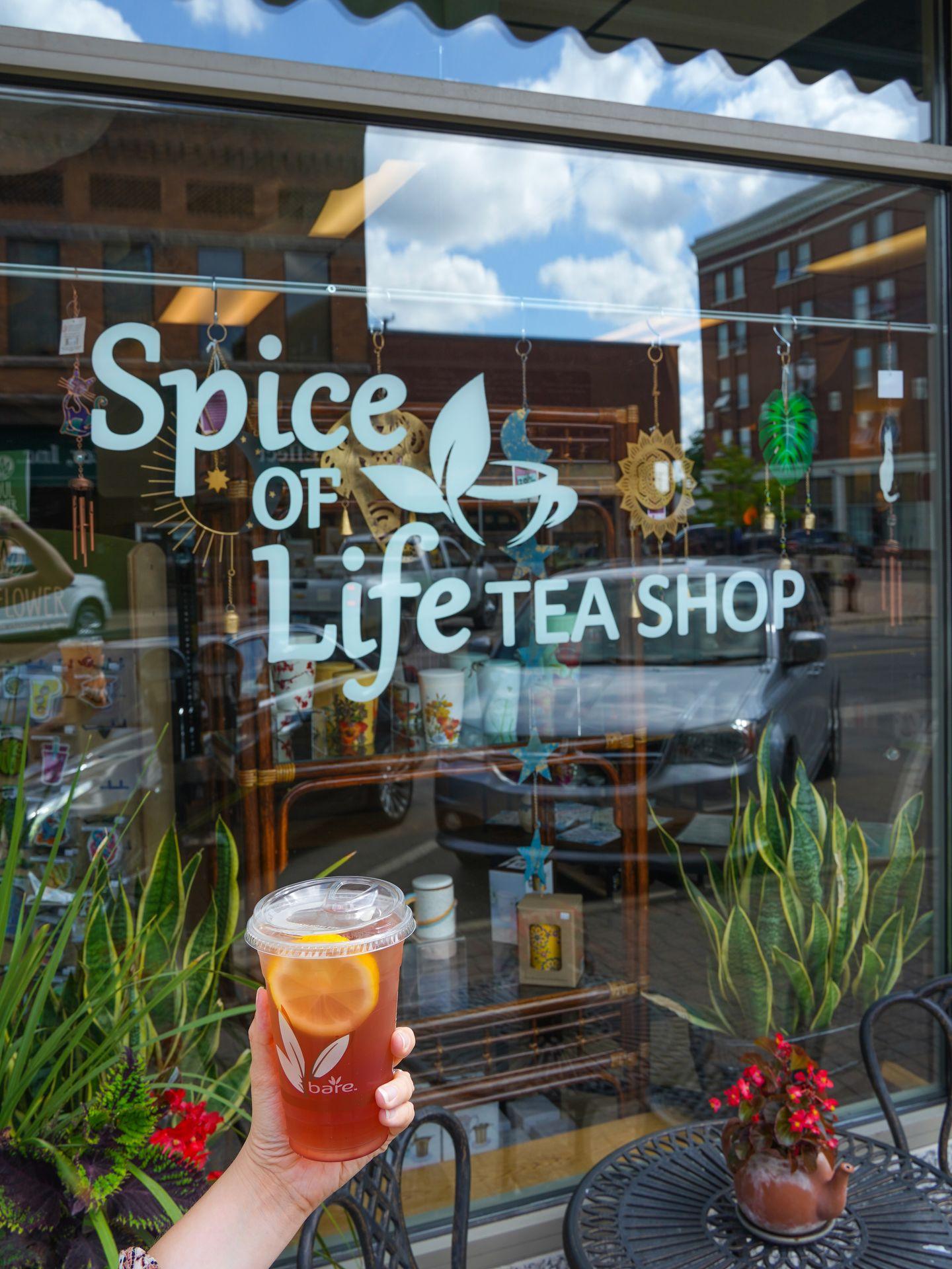 A hand holding a cup of tea in front of Spice of Life Tea Shop