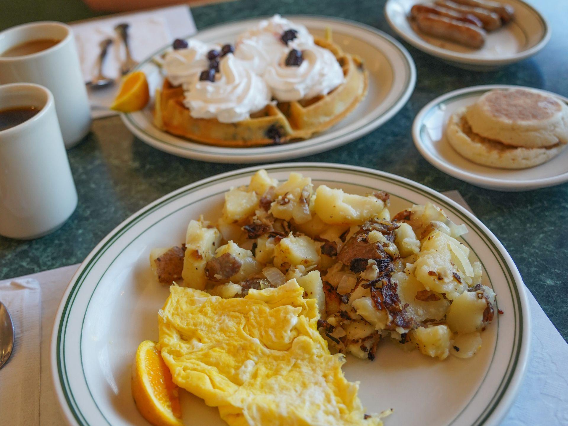 A close up view of a plate with hash browns, scrambled eggs and an orange slice. In the background, there is a waffle with whipped cream, an english muffin and sausage links.