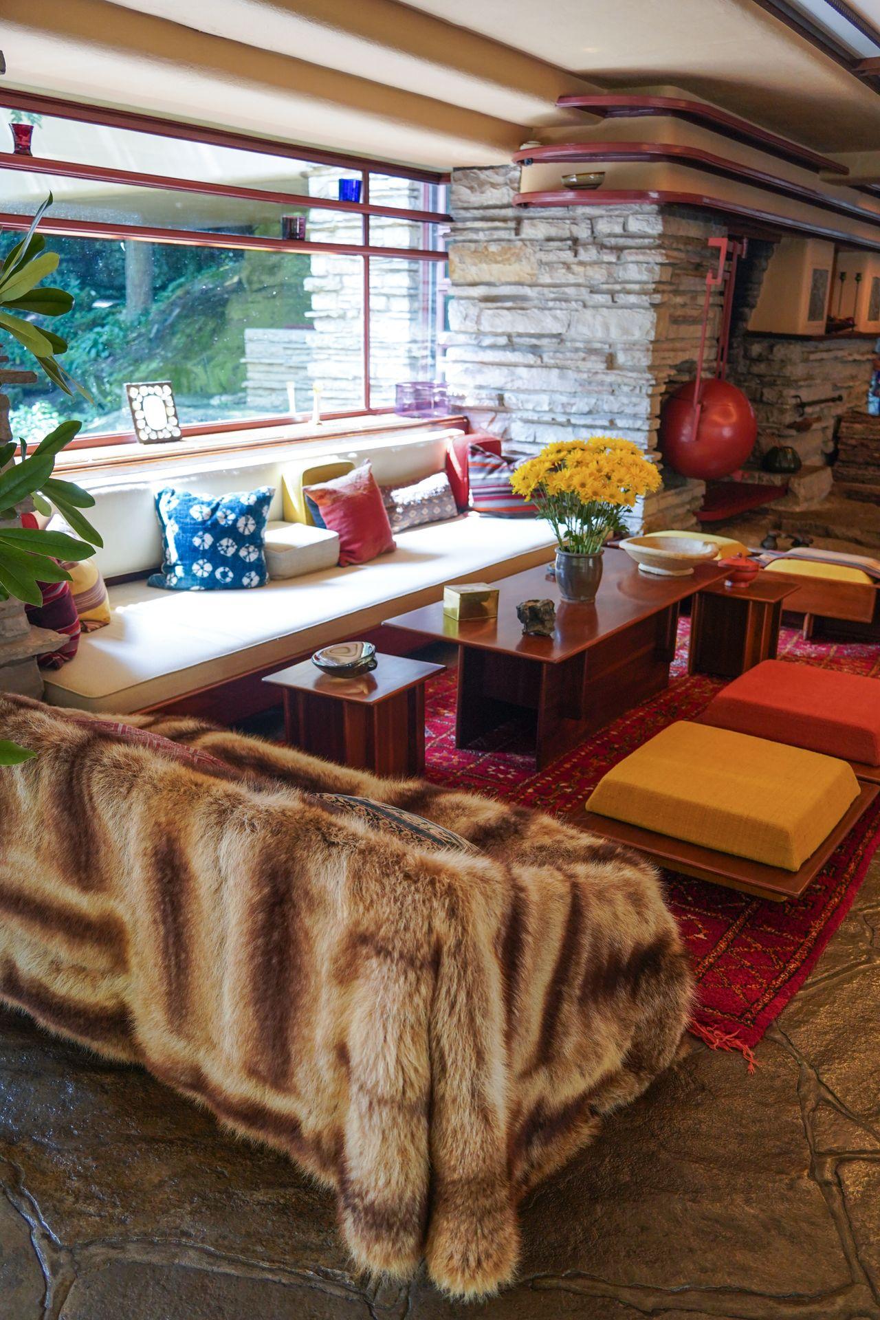 The main living space in Fallingwater. There is a couch against the wall, a fur blanket covering a smaller couch, a red rug and a couple of small tables. There is also a vace of yellow flowers and some colorful throw pillows on the couch.