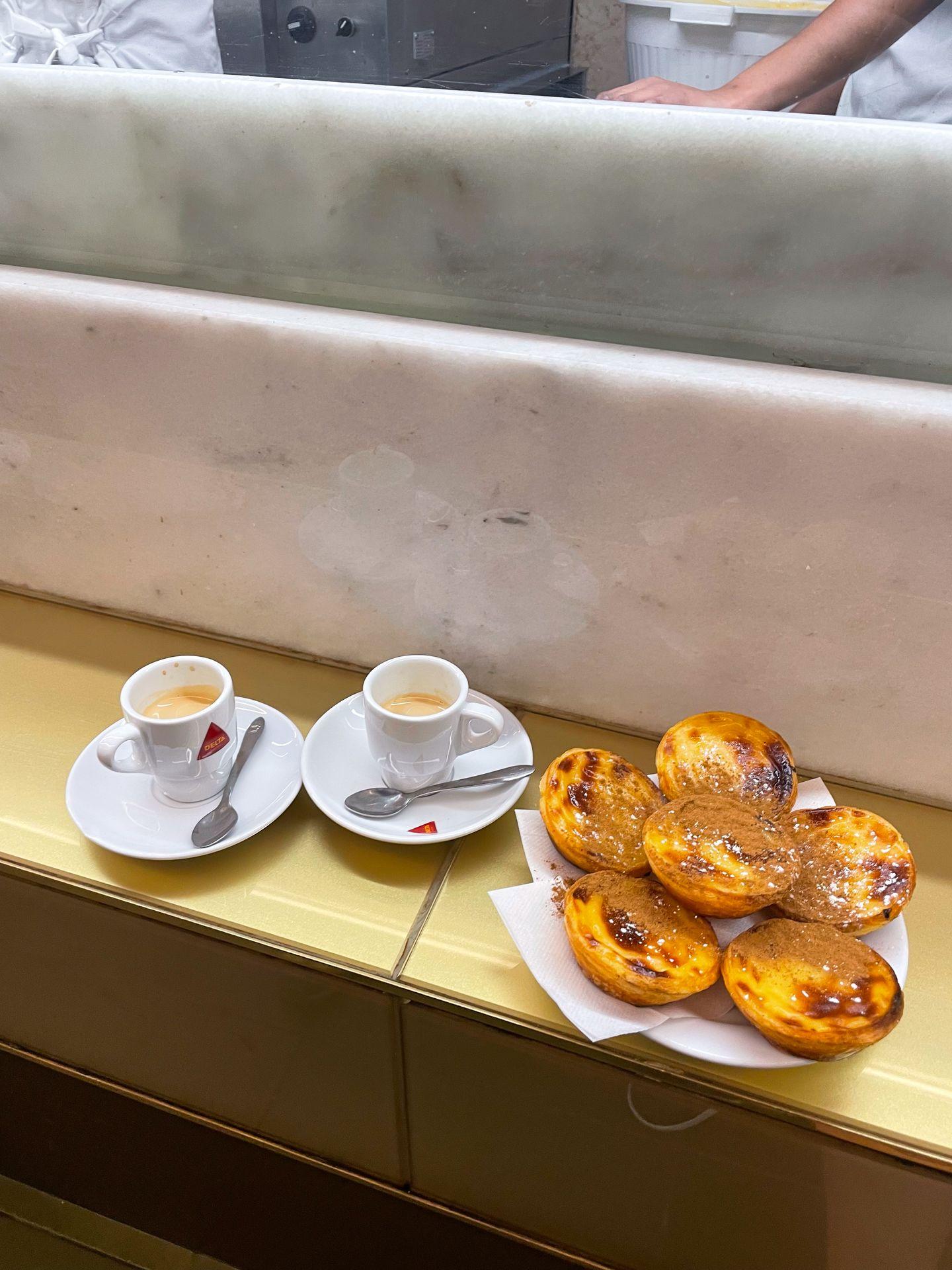 A plate of 6 pastel de natas and two espresso drinks at a bar