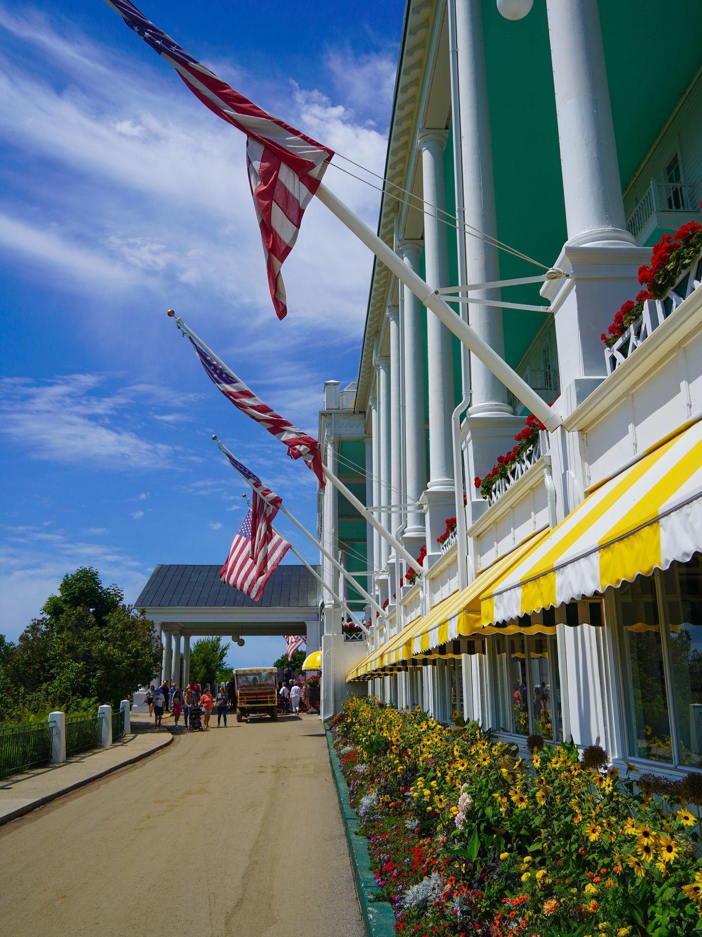 A view of the front of the Grand Hotel. There are white columns, an aqua-colored ceiling of the porch, awnings with yellow and white stripes and flower beds.