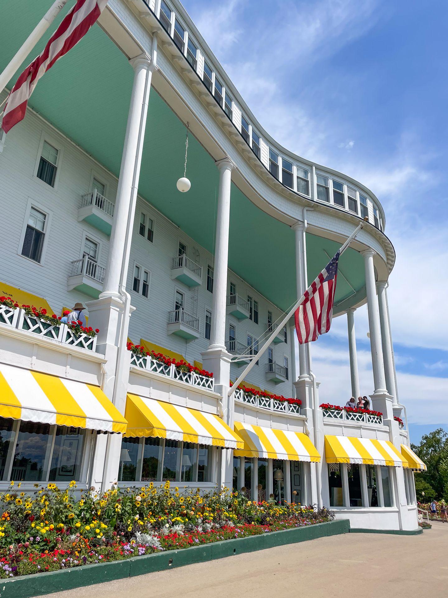 The front porch of the Grand Hotel. It's a white building with columns and yellow and white straped awnings. There are colorful flowers out front and American flags attached to the building.