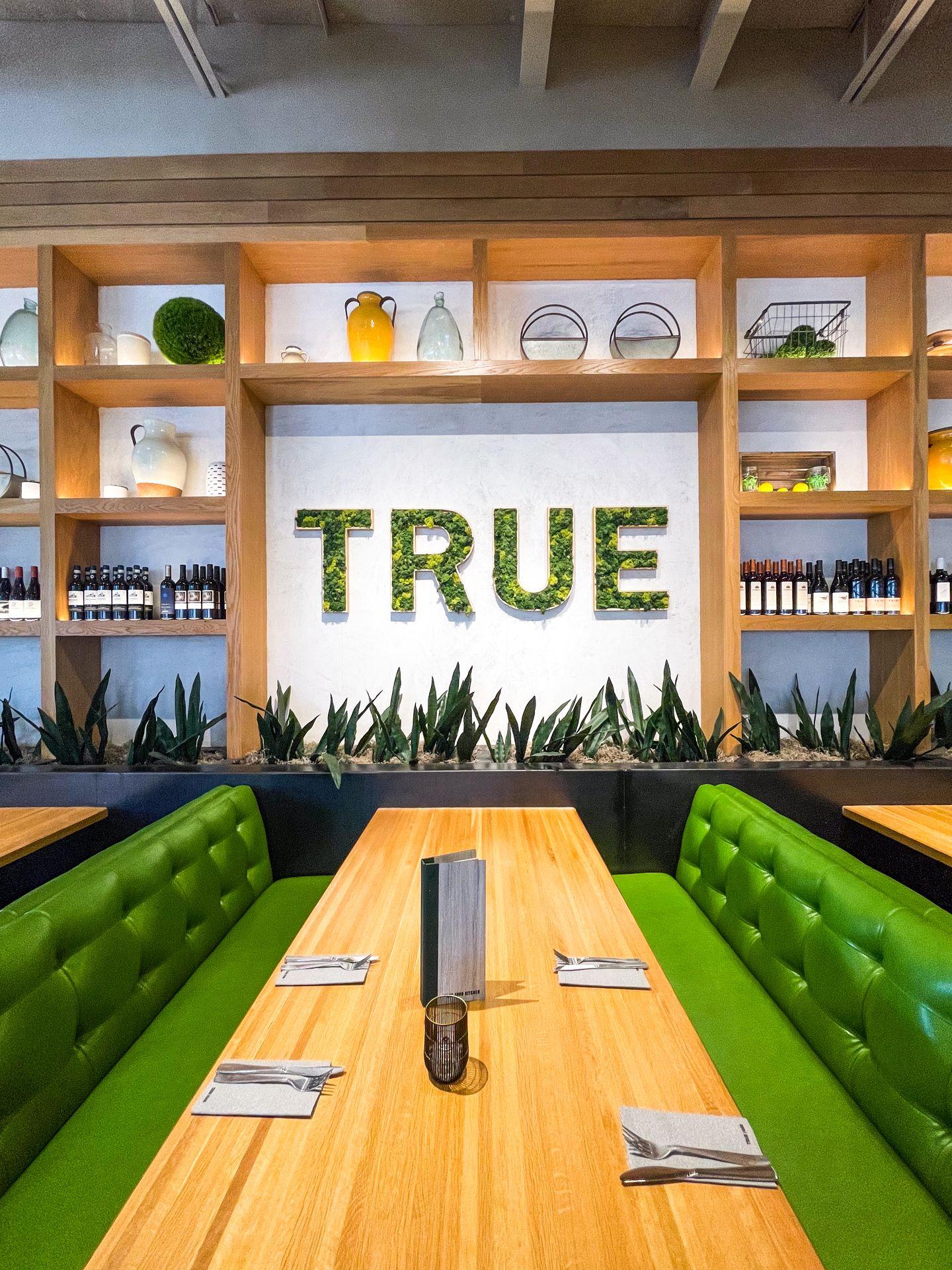 A view of the interior of True Food Kitchen - the word 'true' made of greenery is on a white wall and surrounded by shelving
