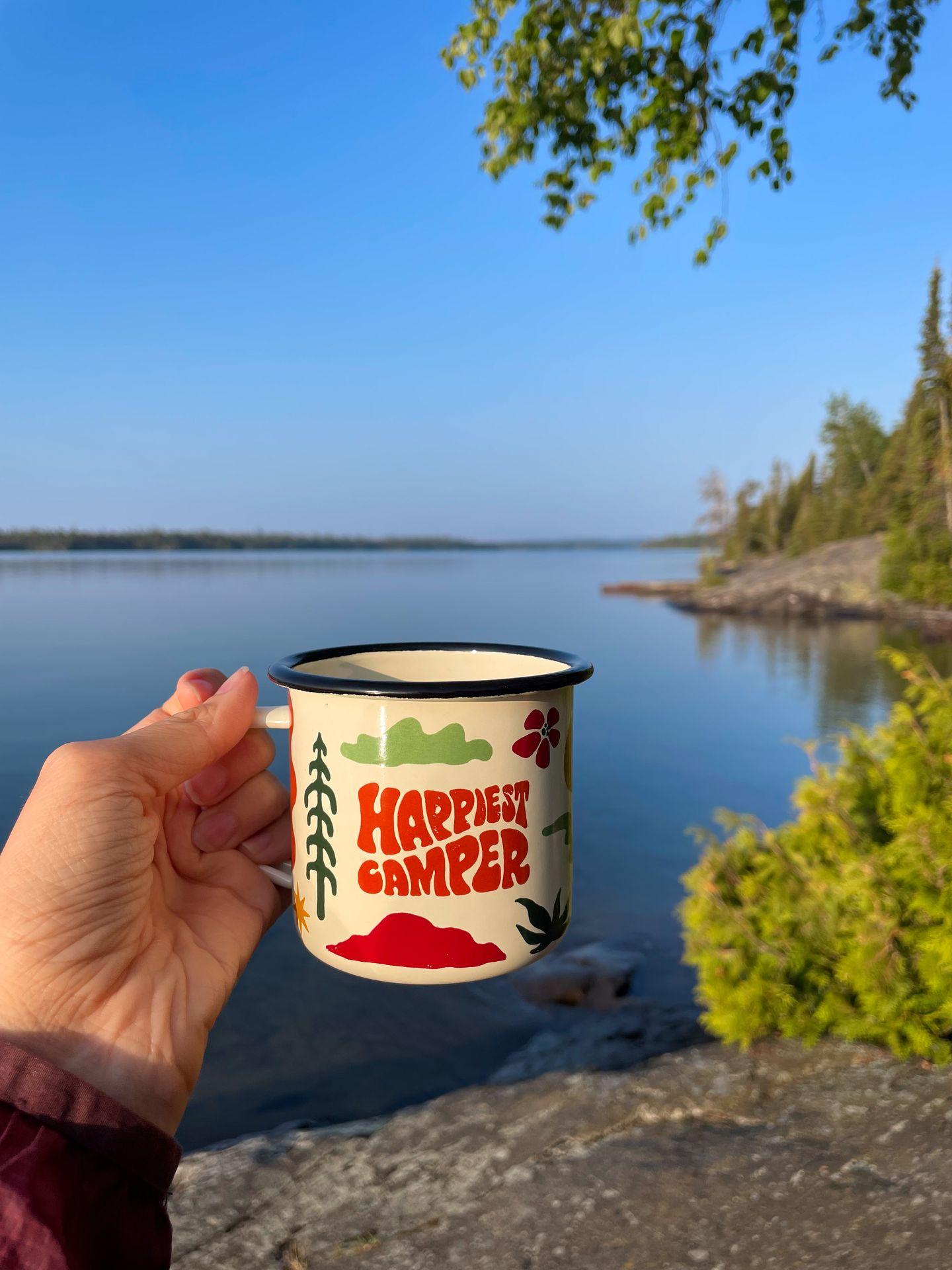 Holding up a mug that reads 'Happiest Camper' on the shores of Lake Superior