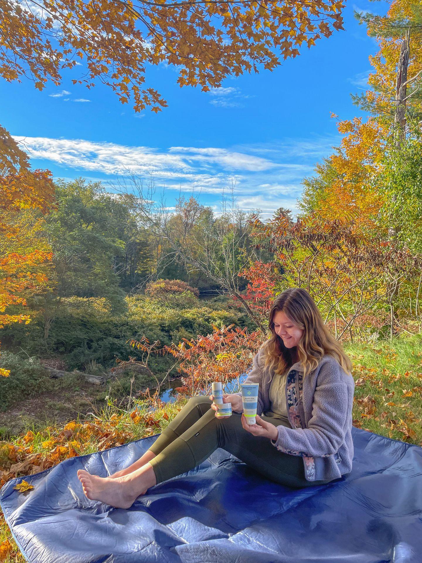 Lydia holding up foot care products from Pedestrian Project while sitting on a blue tarp. There is fall foliage in the background.