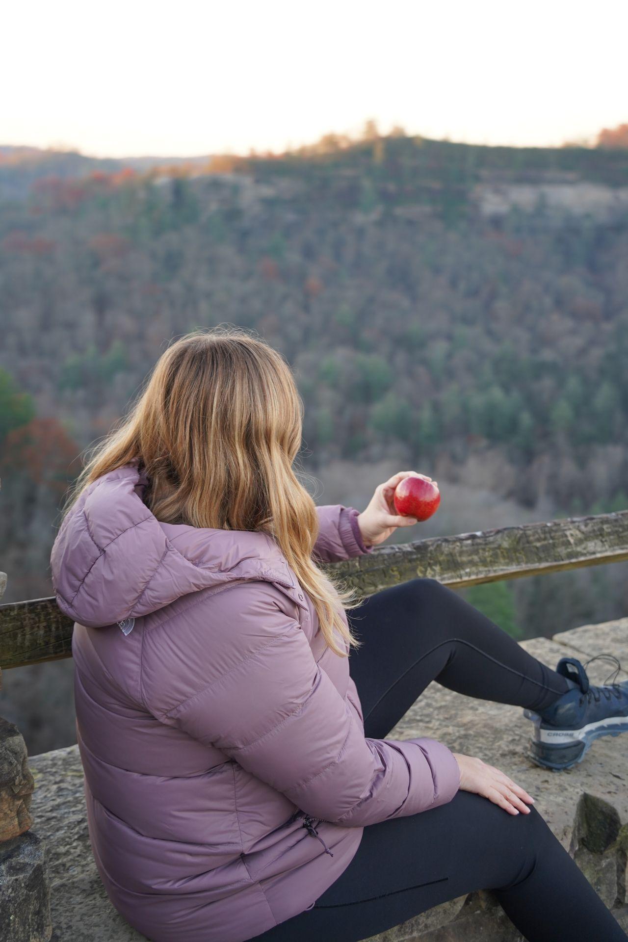 Lydia sits looking at a view of Red River Gorge holding an apple.