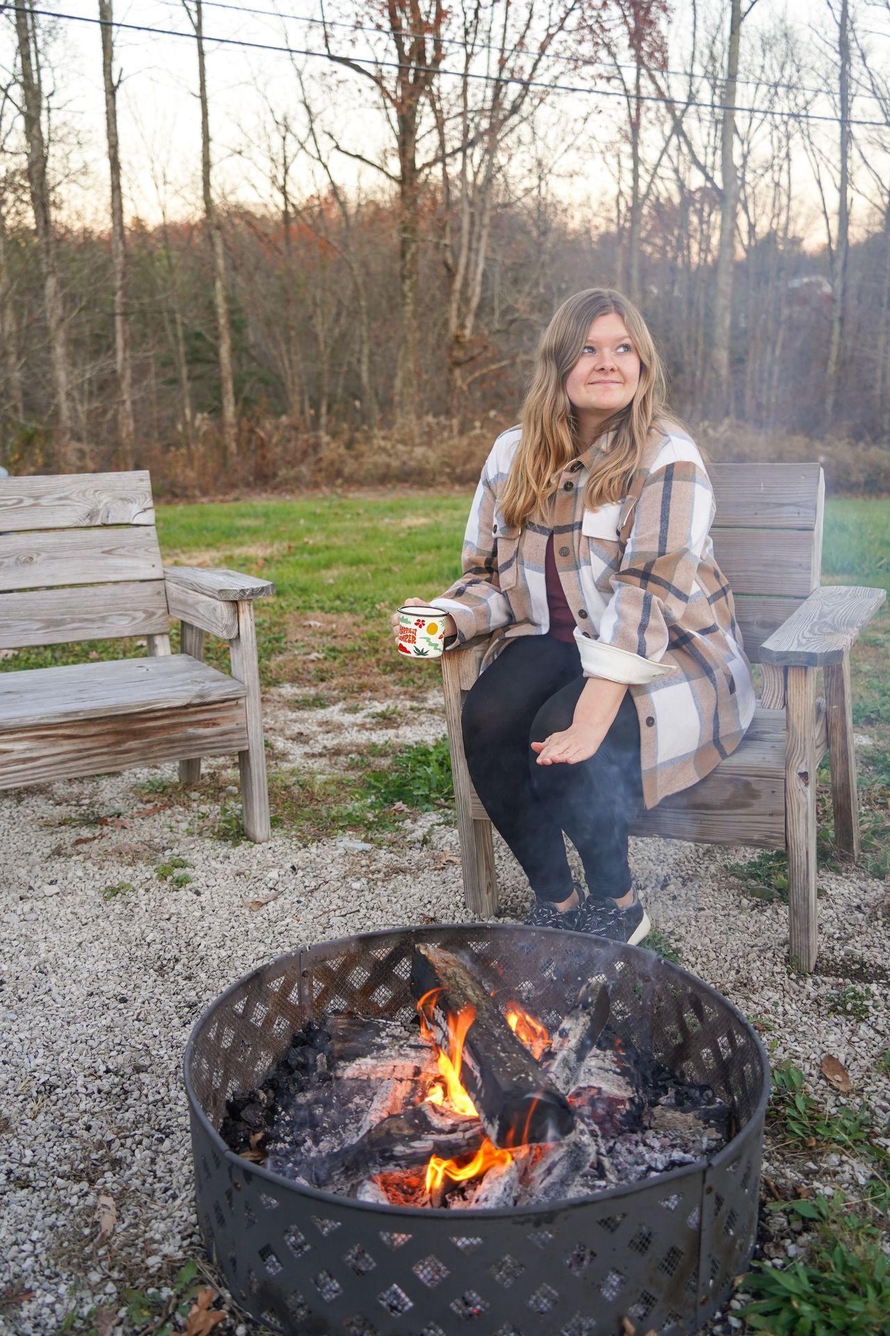 Lydia sitting next to a campfire and warming her hands while holding a mug.