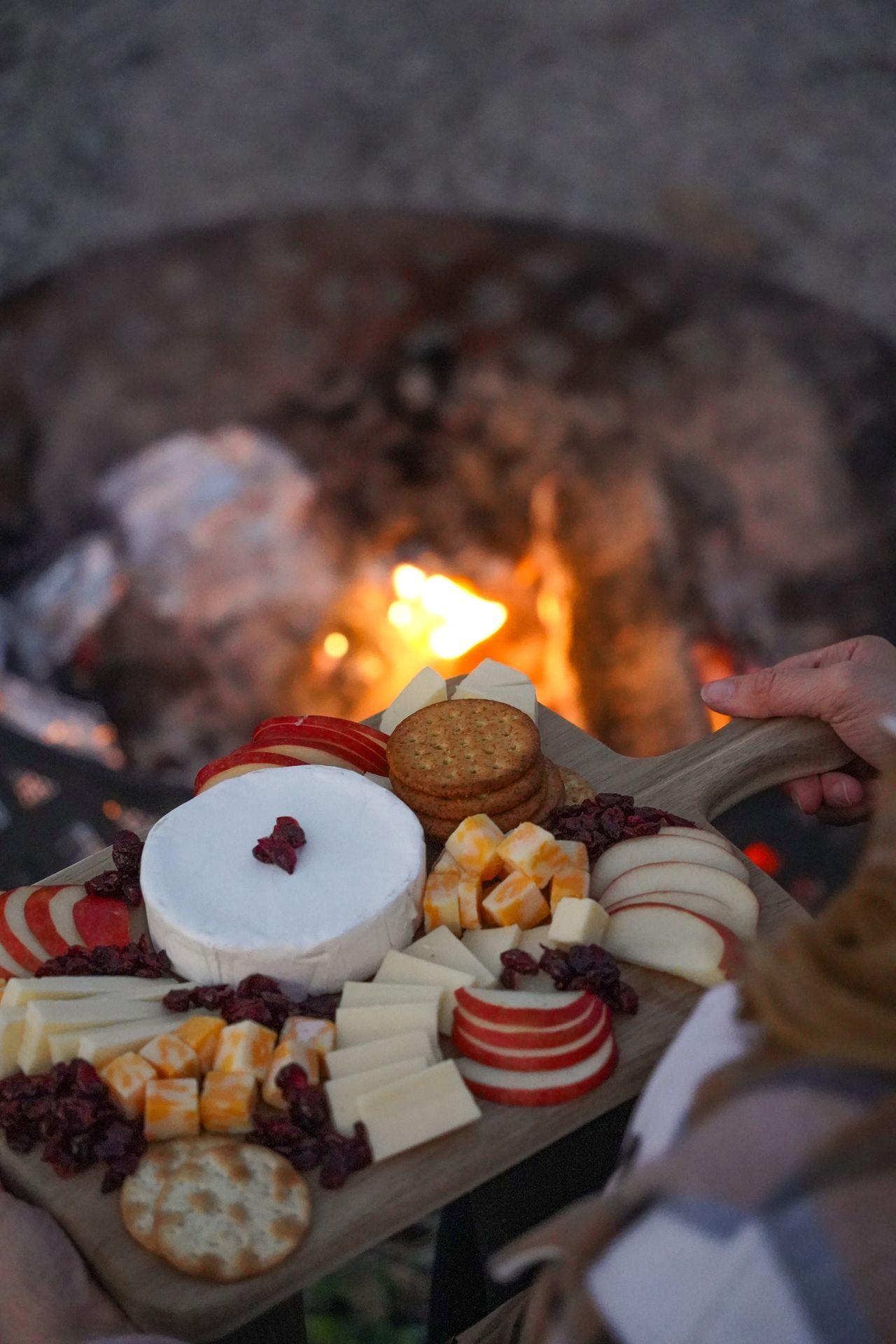Holding up a charcuterie board next to a campfire. The board has 3 cheeses, crackers, apple slices and dried cranberries.
